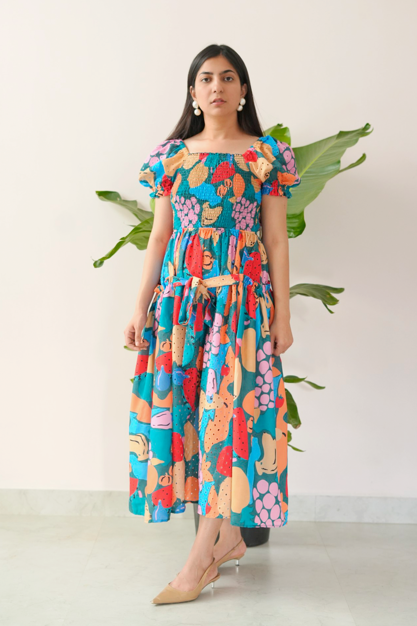Woman wearing a colorful floral dress standing against a white background with green leaves.