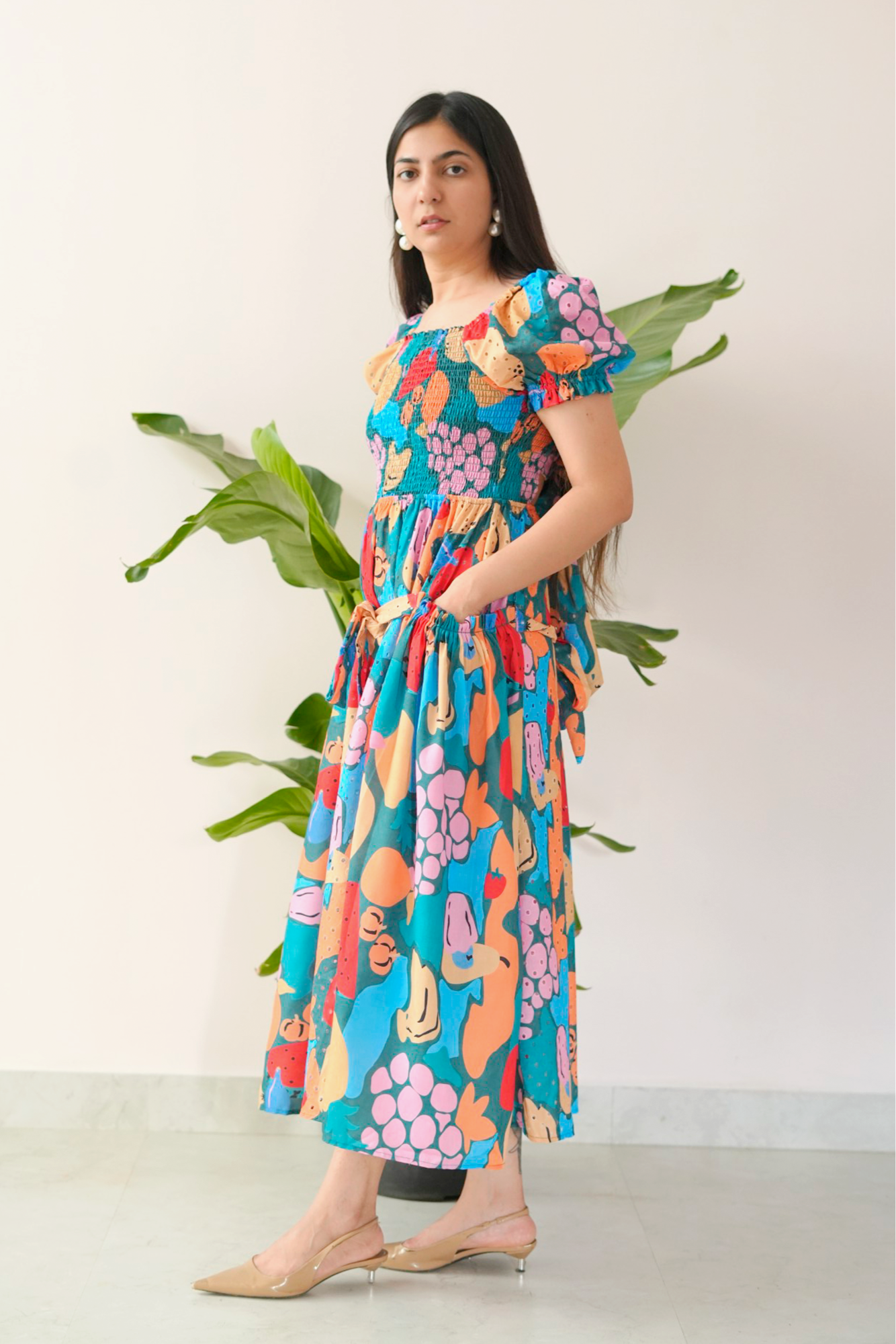 Woman wearing a colorful floral dress standing against a white background with green plants.