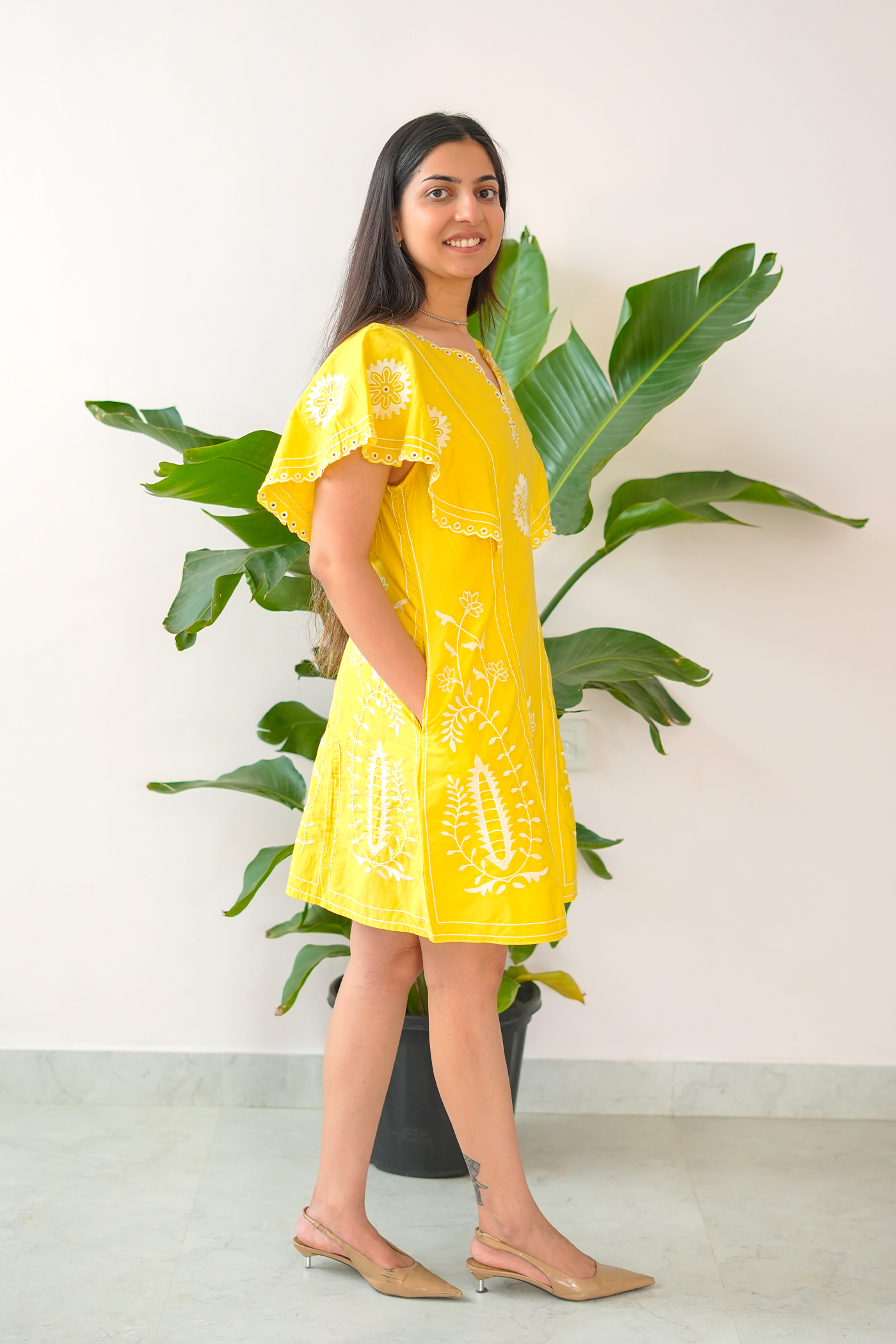 Woman wearing a yellow dress with white patterns standing in front of a plant.