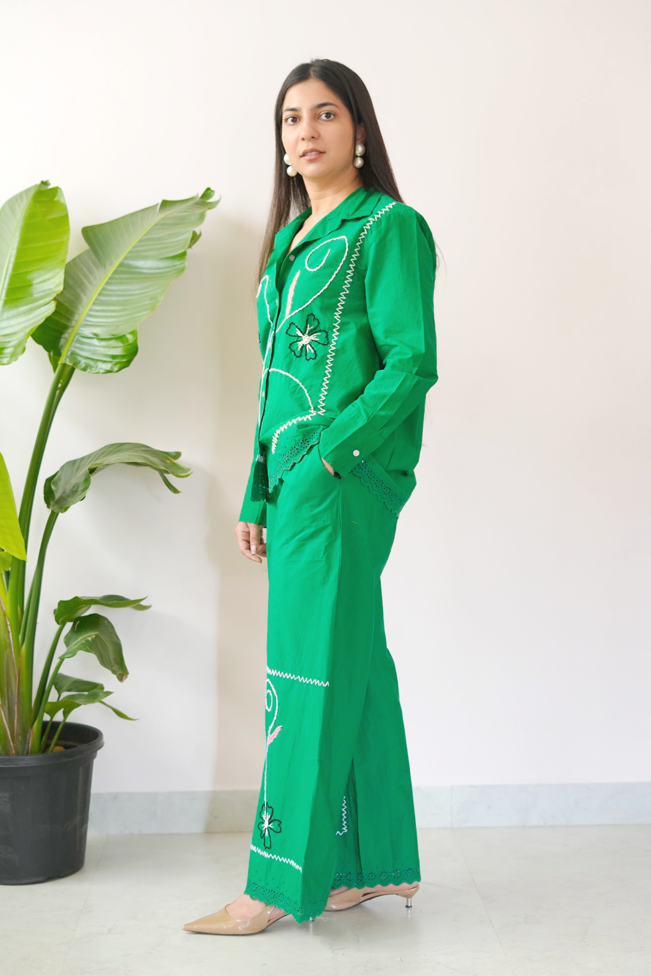 Woman wearing a green outfit with white patterns standing next to a potted plant.