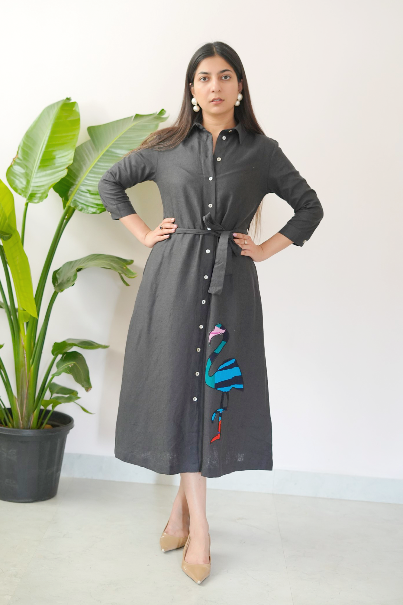 Woman wearing a gray dress with floral embroidery in front of a white wall and plant.