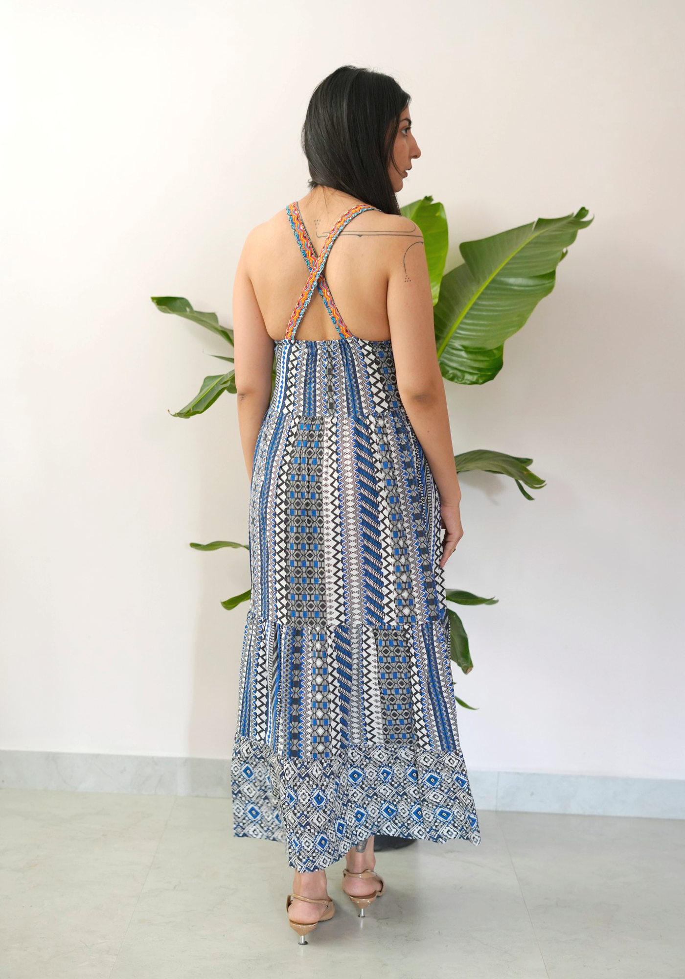 Woman in a patterned dress standing against a white wall with a plant.