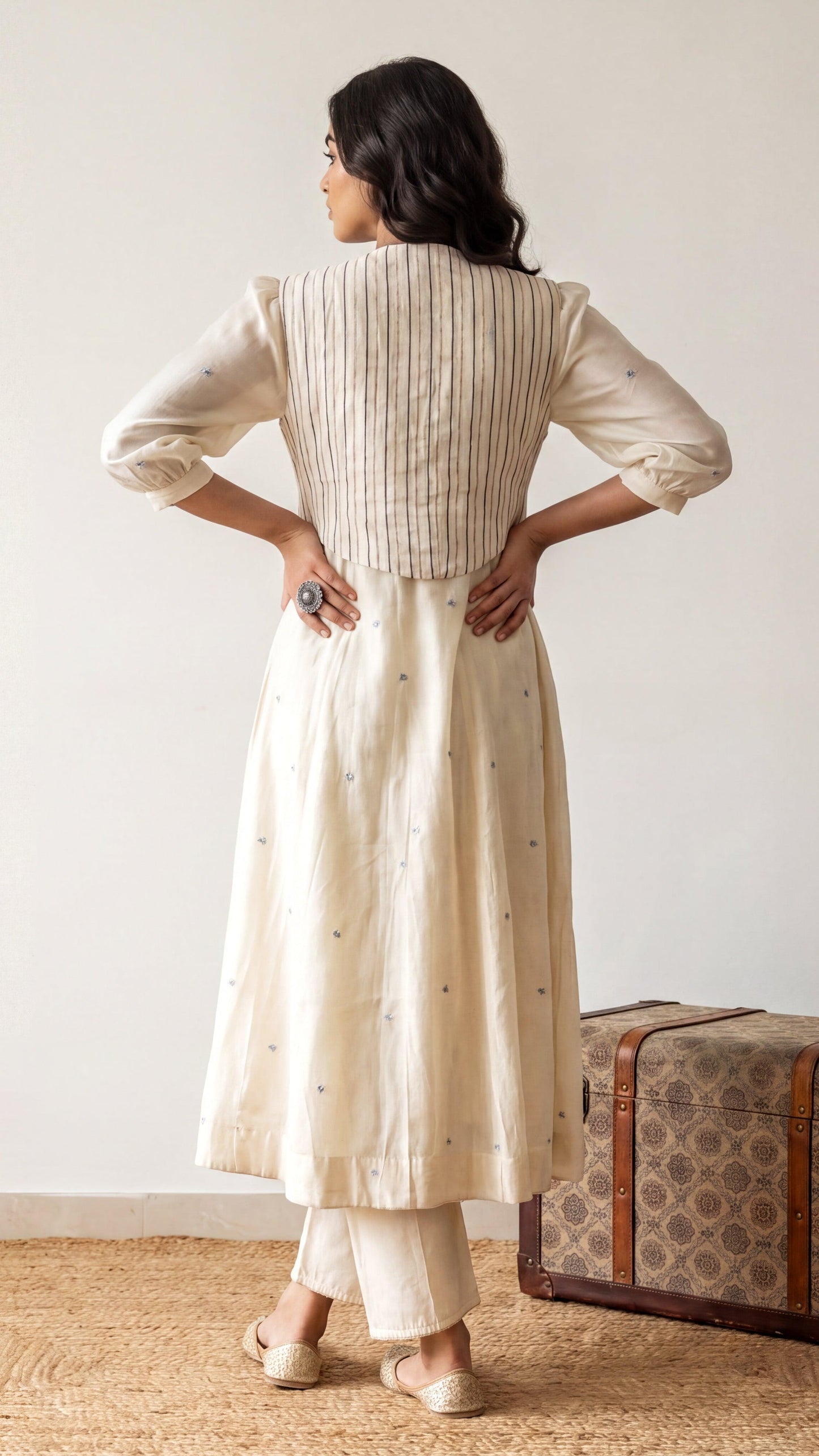 Woman wearing a long, light-colored dress standing next to a patterned suitcase against a plain background.