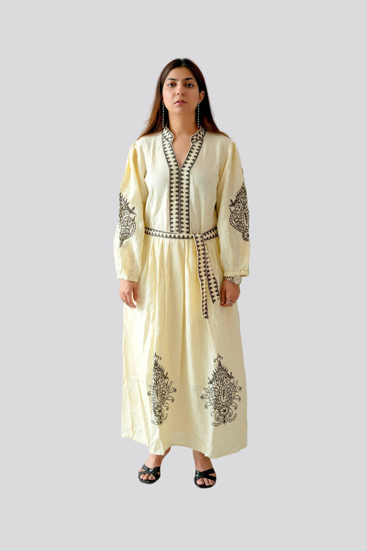 Woman wearing a cream-colored dress with black embroidery standing against a white wall with green leaves.