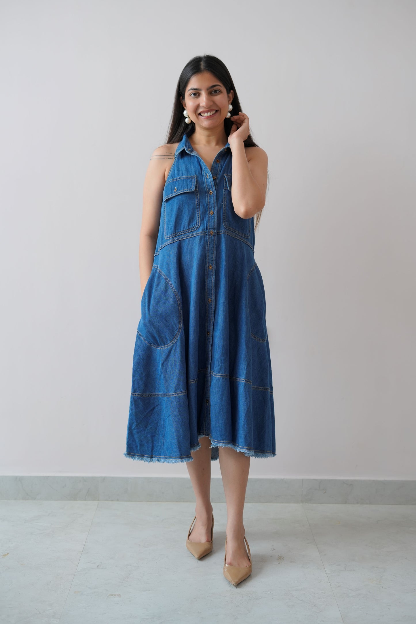 A woman standing in a room with a plain white wall, wearing a blue denim sleeveless dress with a classic collar and side pockets.