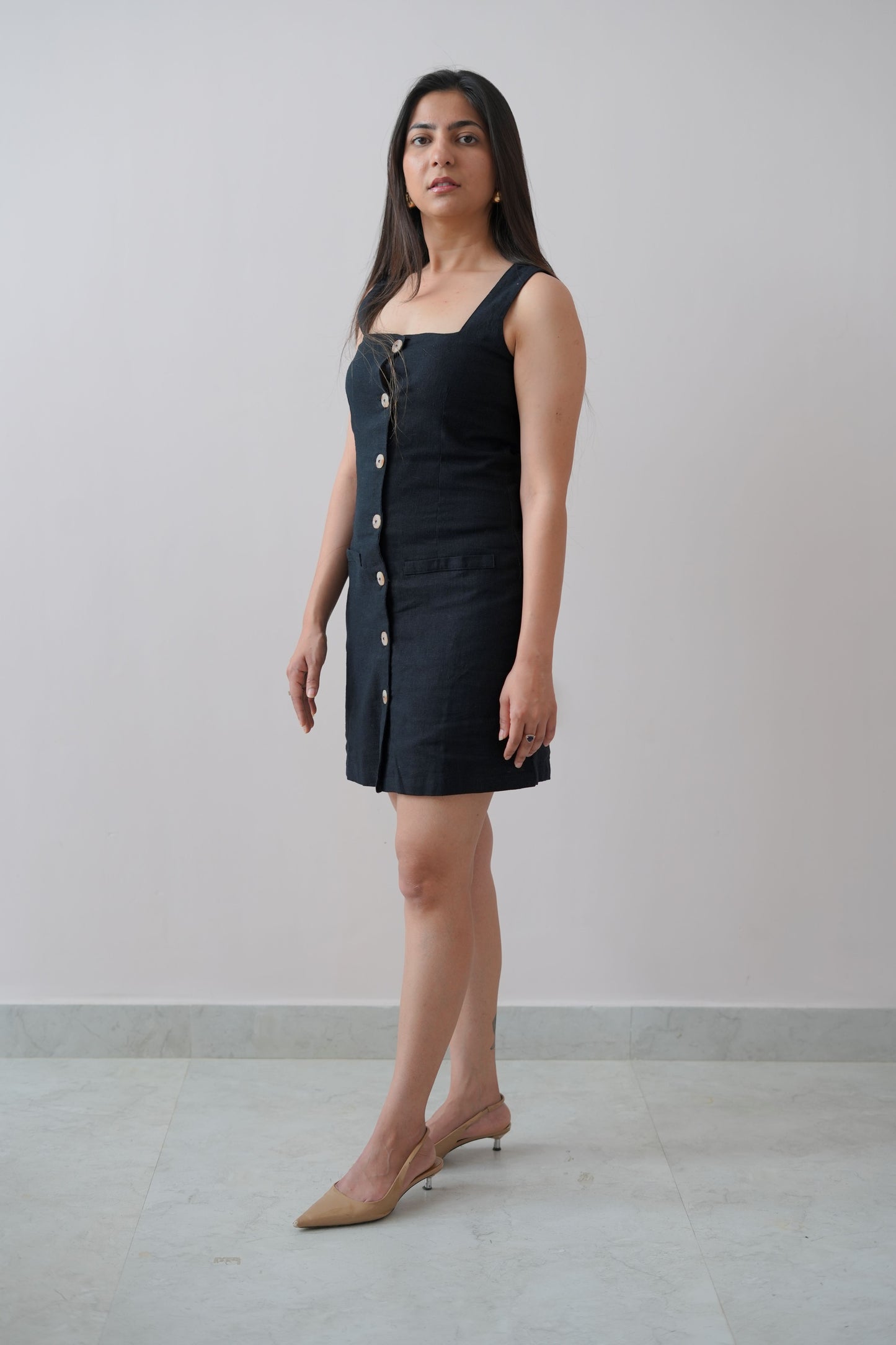 A woman standing in a studio setting, wearing a beige linen short sleeveless dress with square neckline and vertical button details.