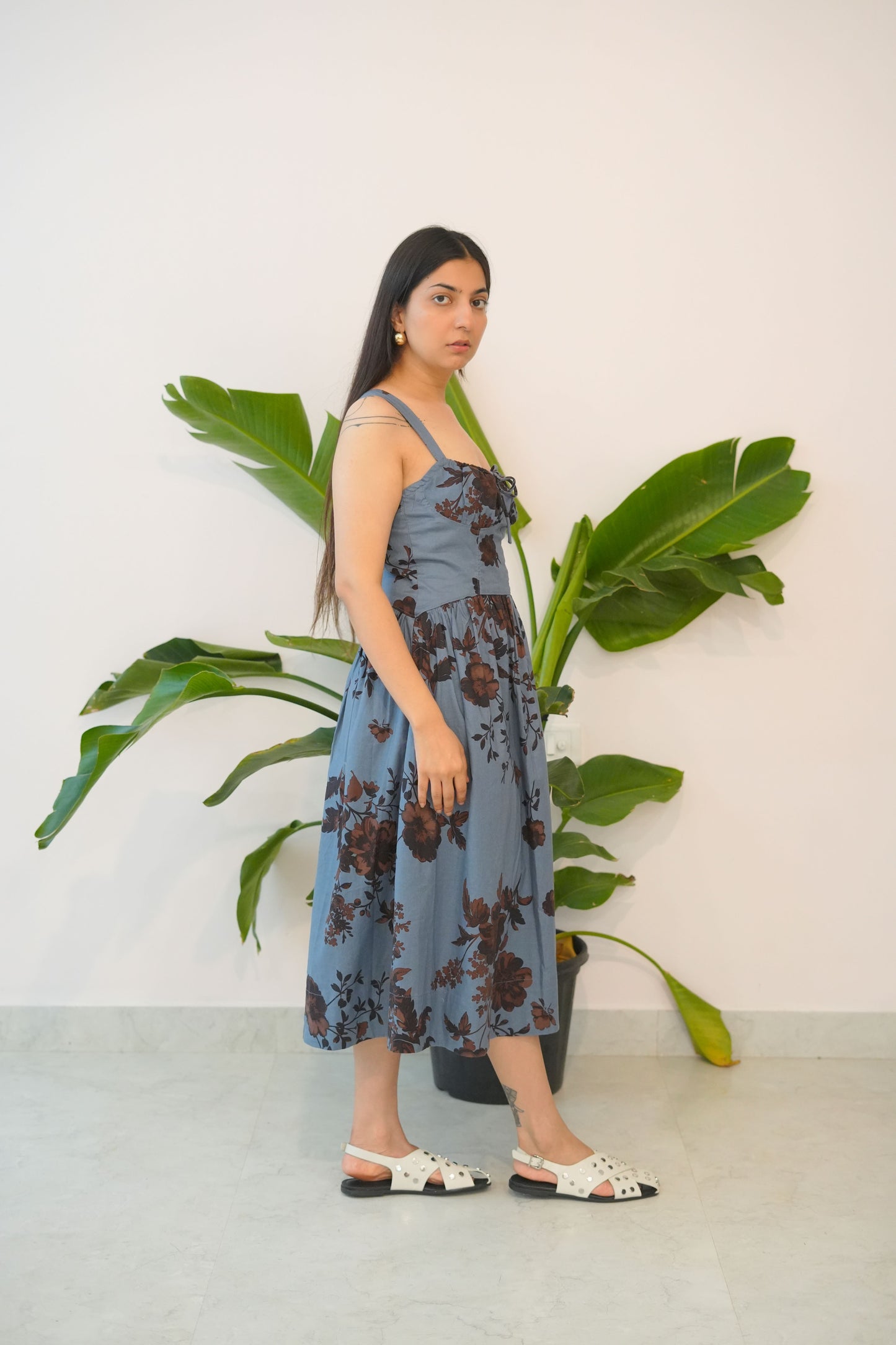 Woman in a blue floral dress standing next to a large green plant indoors.