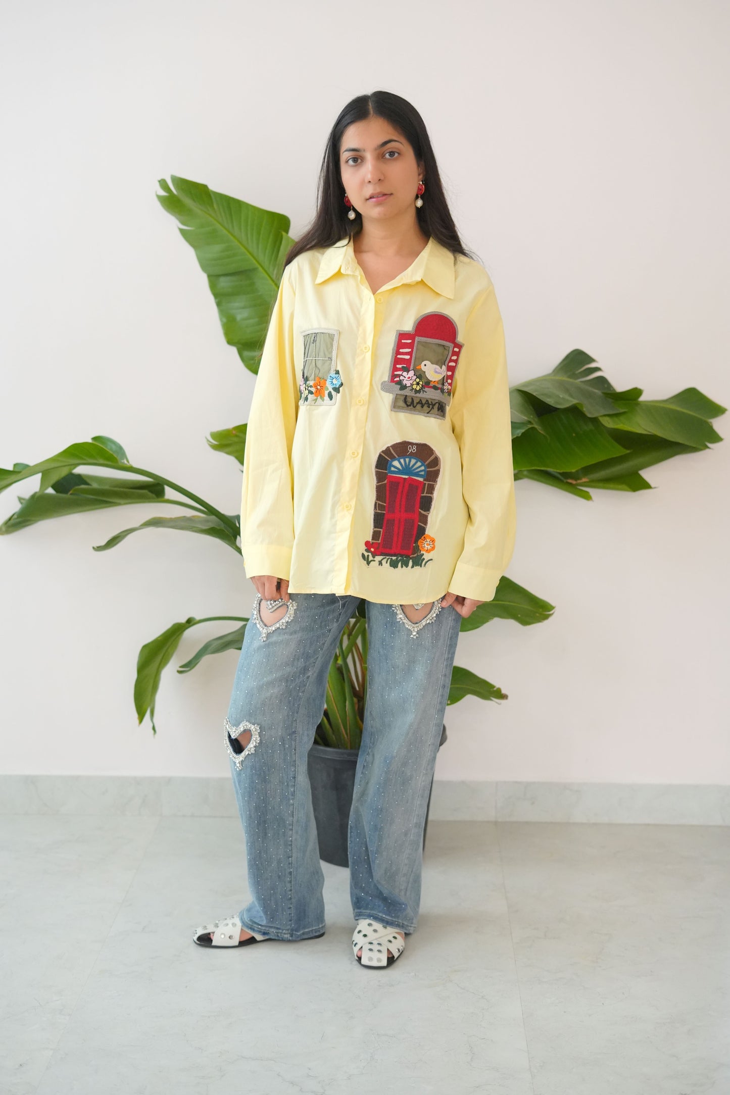 Person wearing a yellow sweatshirt with graphic designs and blue jeans standing in front of green plants.