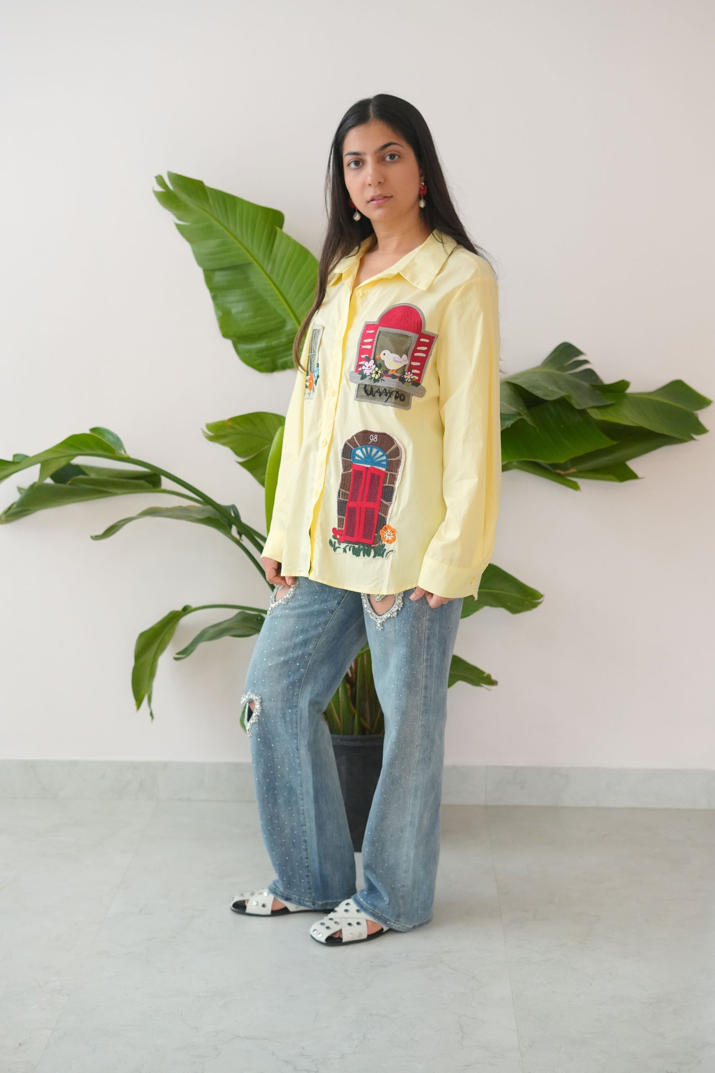 Person wearing a yellow sweatshirt with a graphic design and blue jeans, standing against a white wall with green plants.
