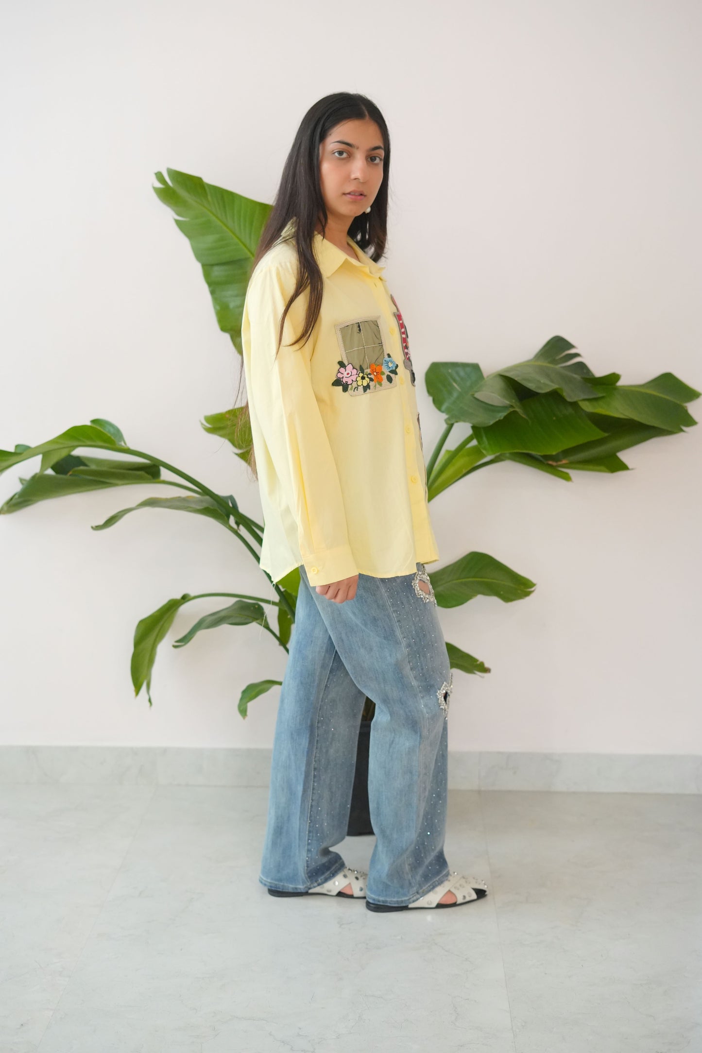 Person wearing a yellow sweatshirt and blue jeans standing next to a plant indoors.