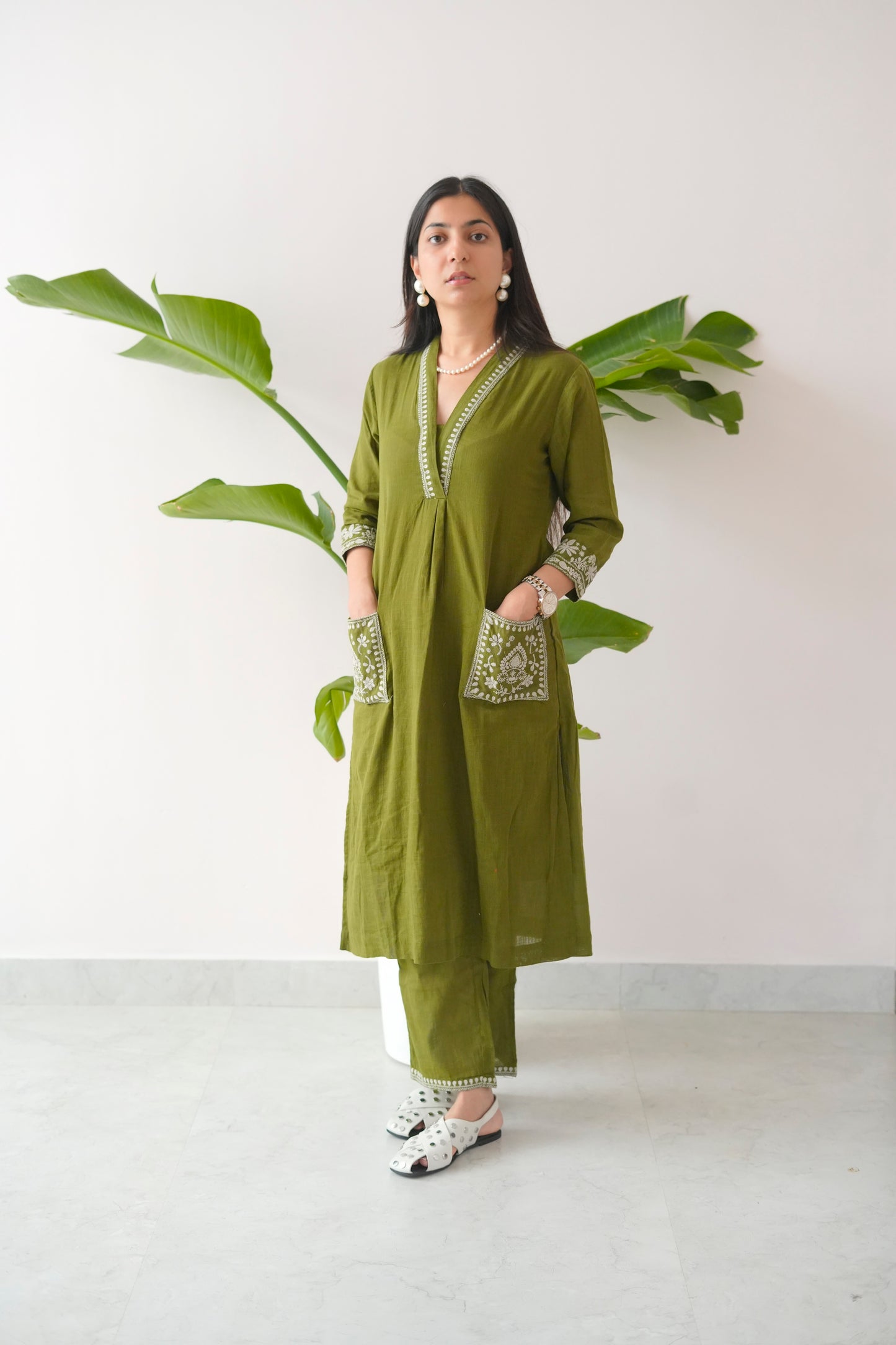 Woman wearing a green traditional outfit with white patterns, standing against a white wall with a plant.