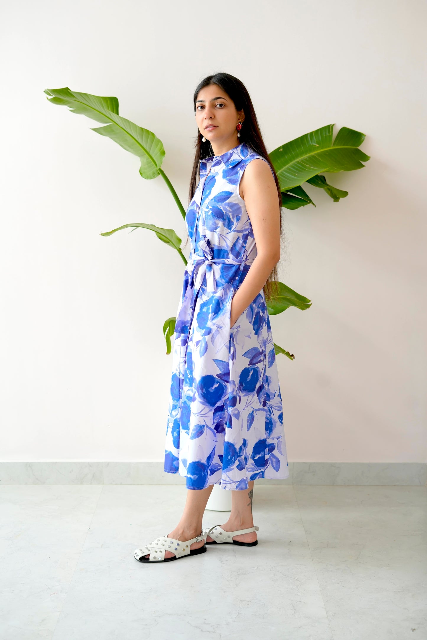 Woman wearing a blue floral dress standing next to a plant against a white wall.