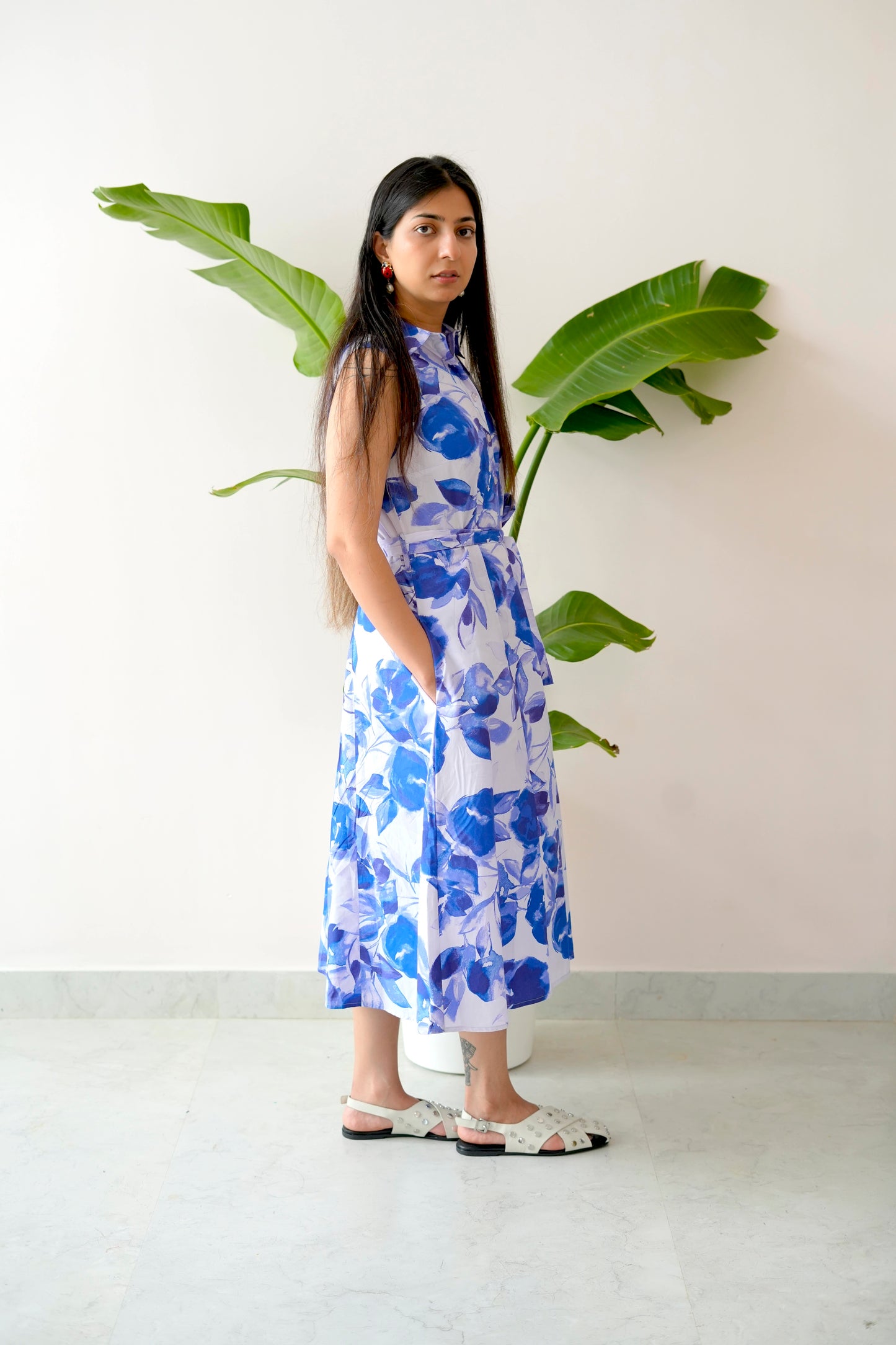 Woman in a blue floral dress standing next to a plant against a white wall.