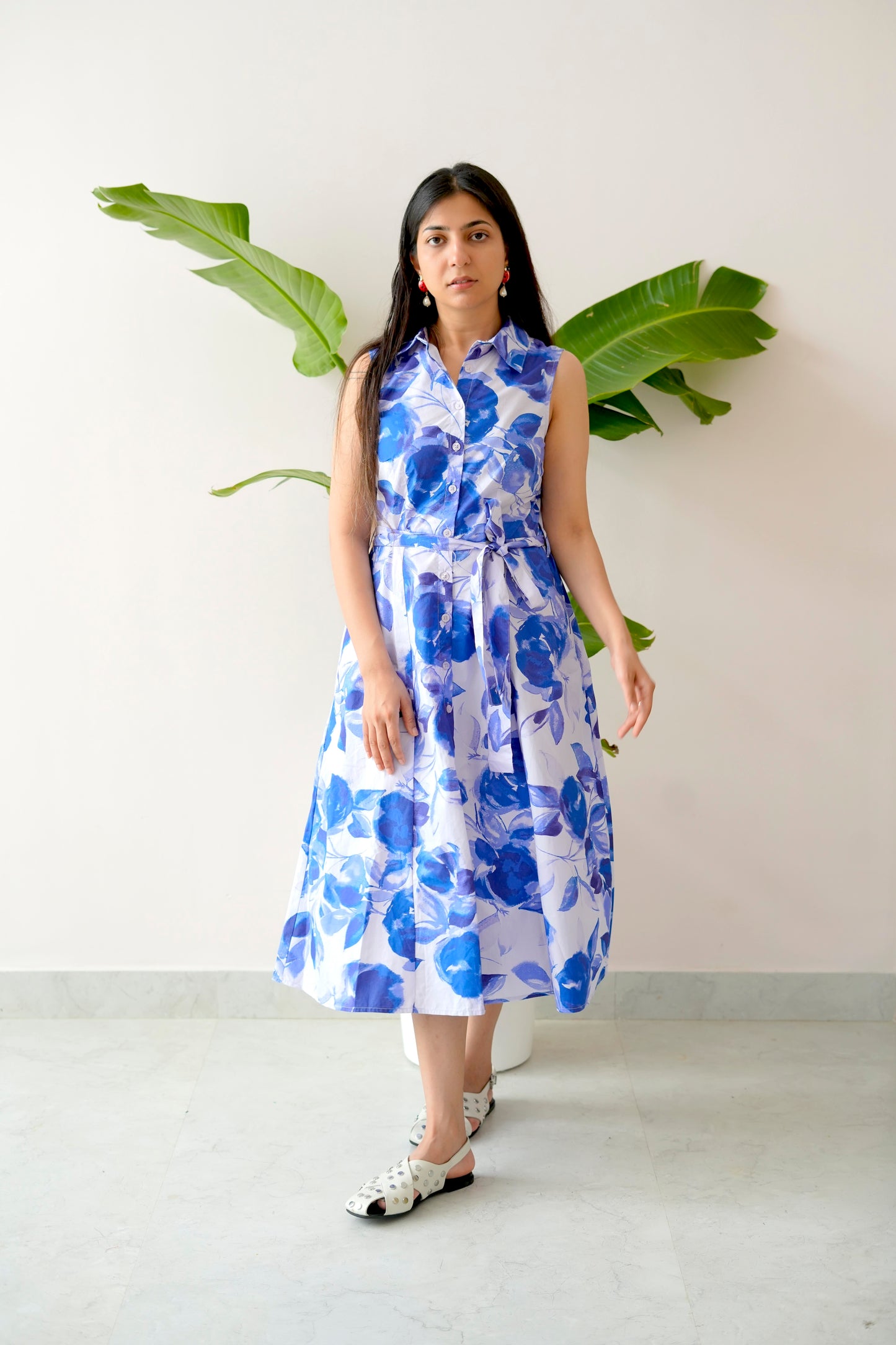 Woman wearing a blue floral dress standing against a white wall with green leaves.