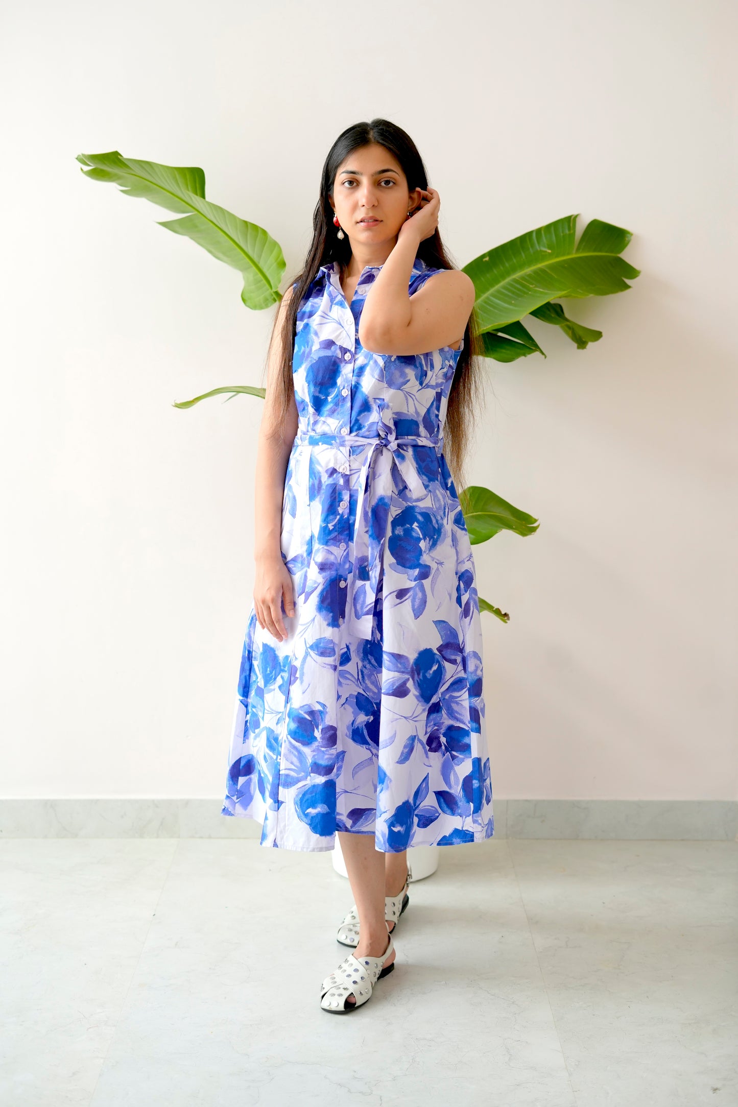 Woman wearing a blue floral dress standing against a white wall with green leaves.