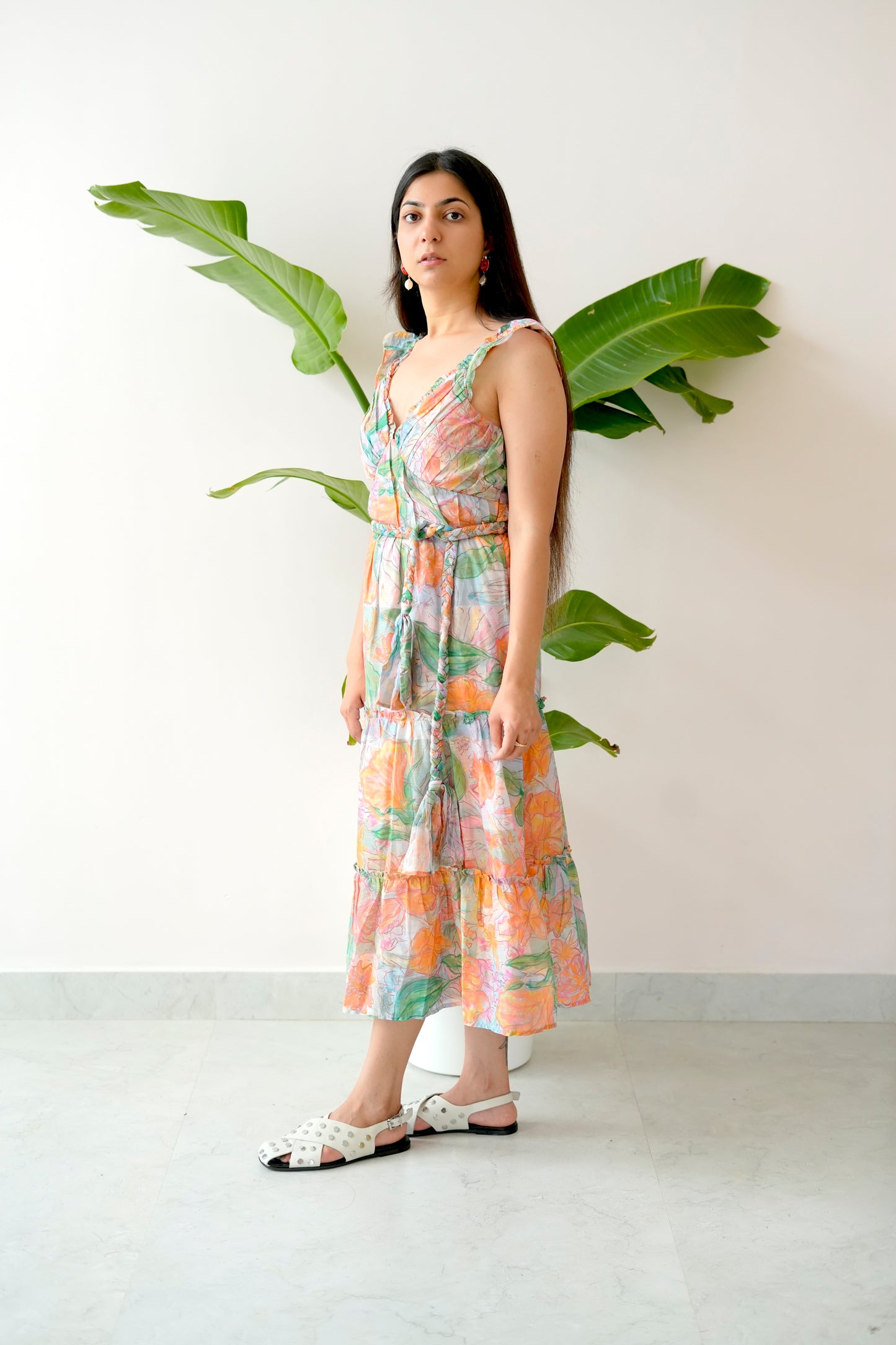 Woman wearing a floral dress standing next to a plant against a white wall.