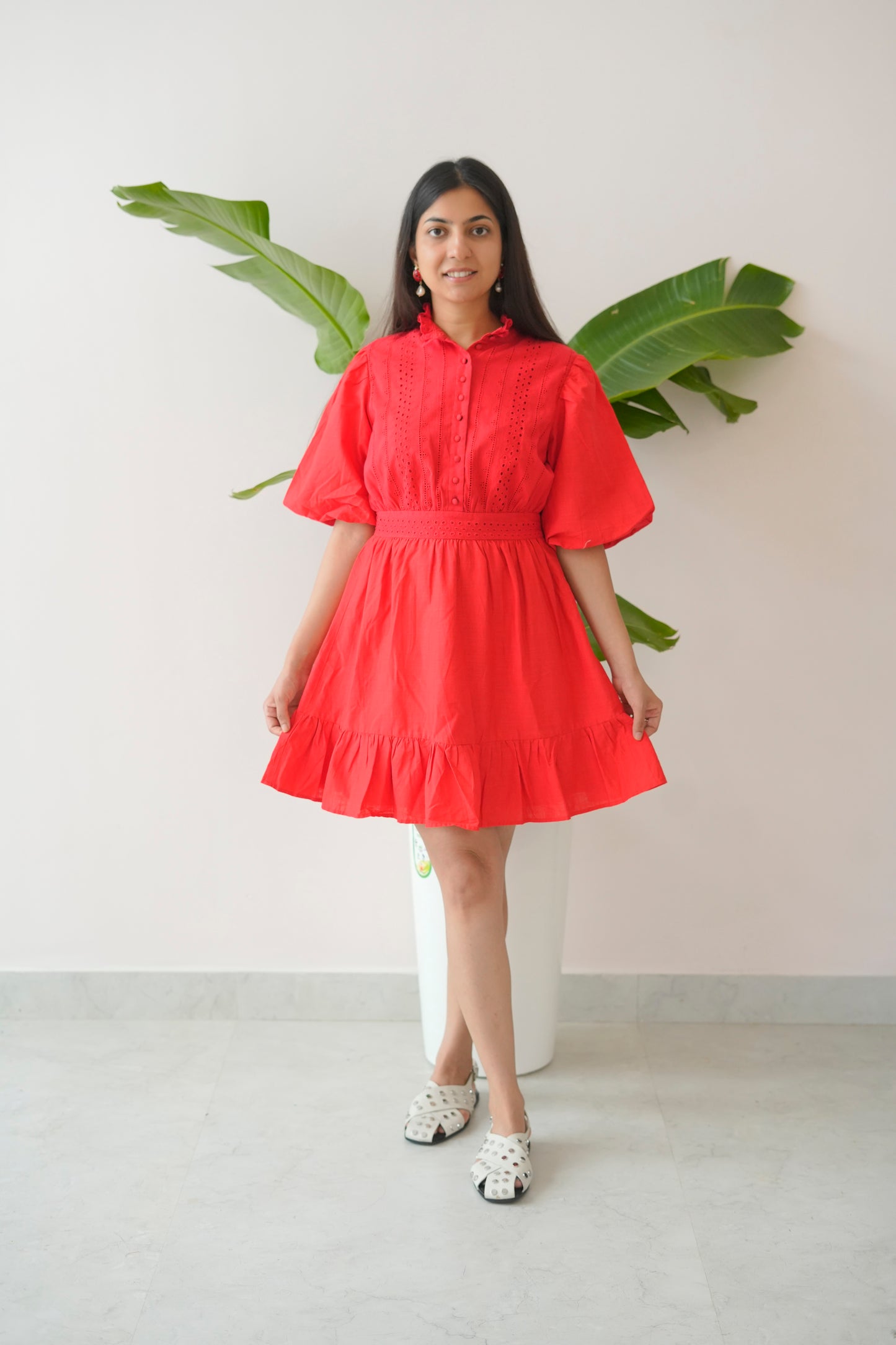 Woman wearing a red dress standing against a white wall with green leaves.