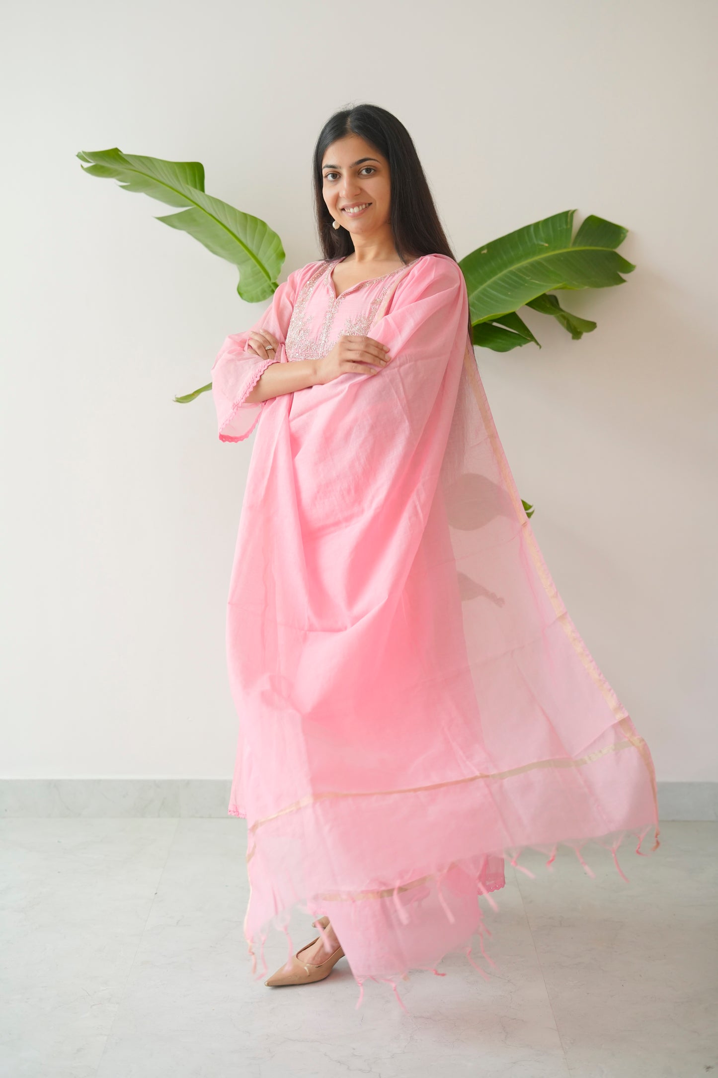 Woman in a pink traditional outfit standing against a white wall with green leaves.