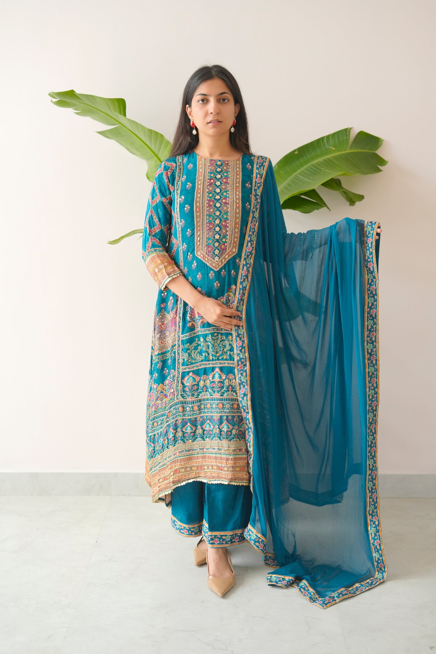 Woman wearing a blue embroidered traditional outfit with a matching dupatta, standing against a white background with green leaves.
