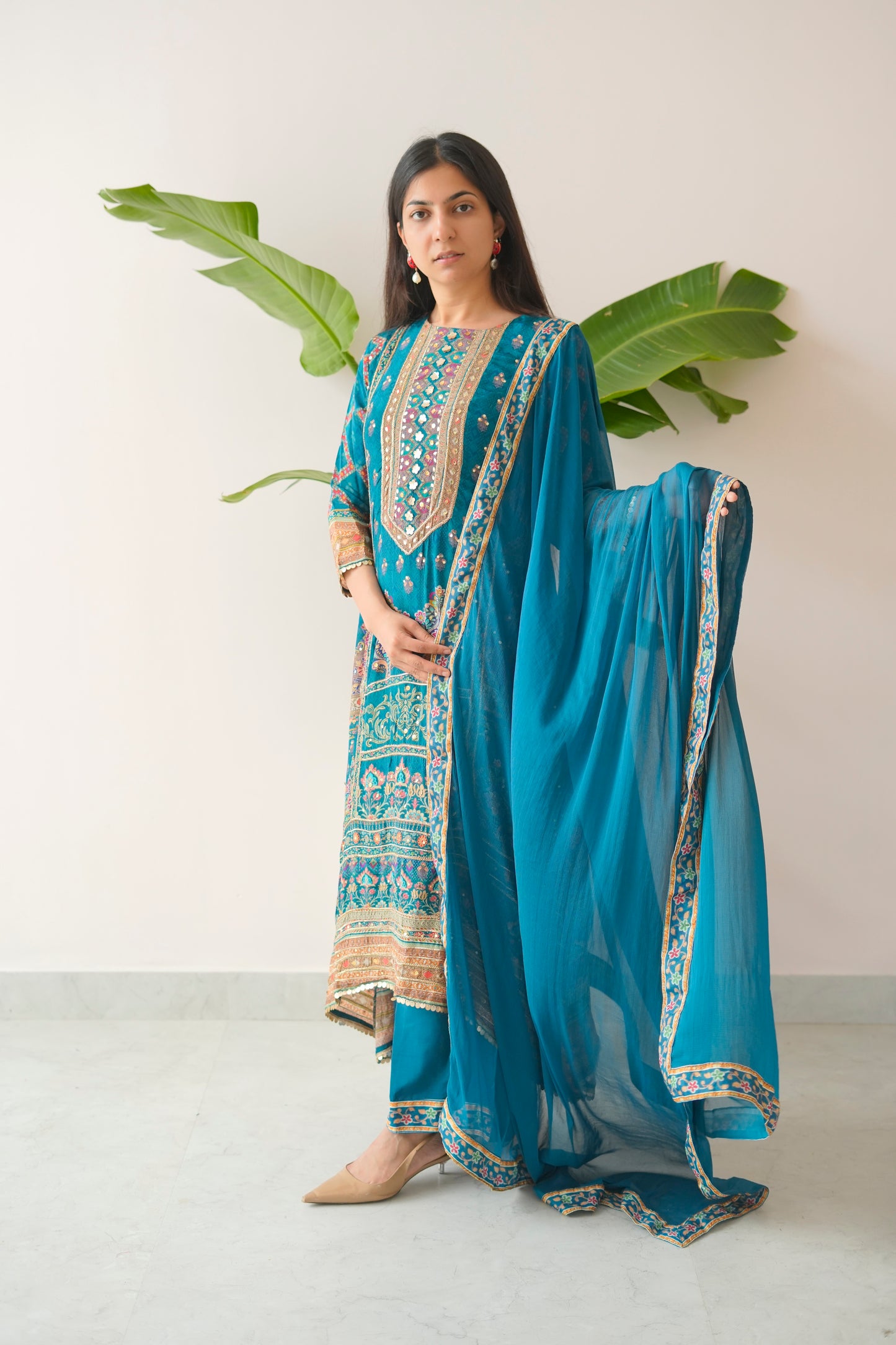 Woman wearing a blue embroidered outfit with a matching dupatta, standing against a white background with green leaves.