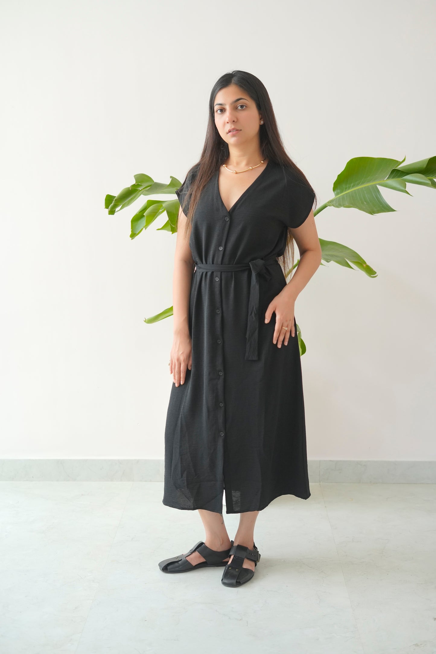Woman wearing a black dress standing in front of green plants against a white wall.
