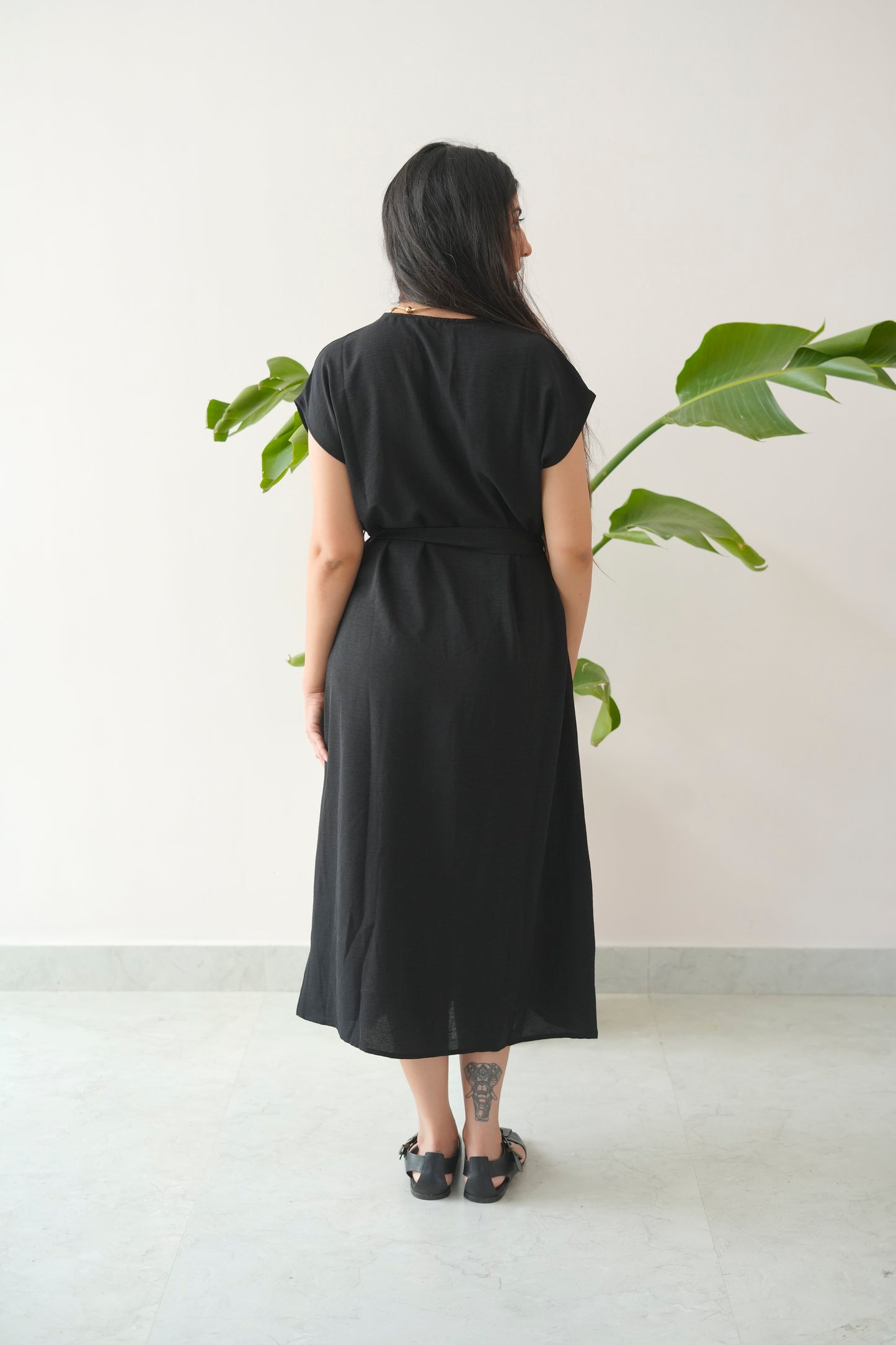 Person wearing a black dress standing in front of a plant against a white wall.