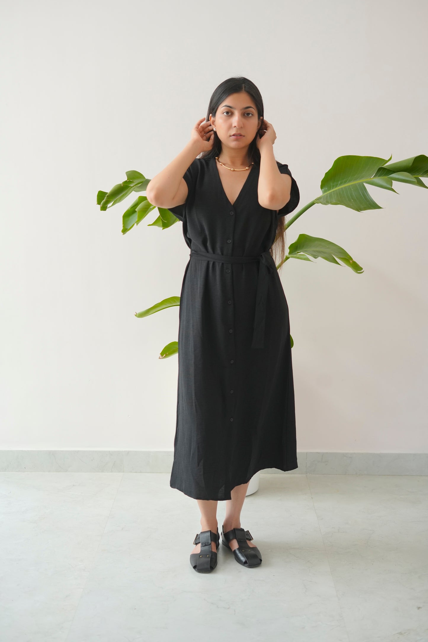 Woman wearing a black dress standing in front of a plant against a white wall.