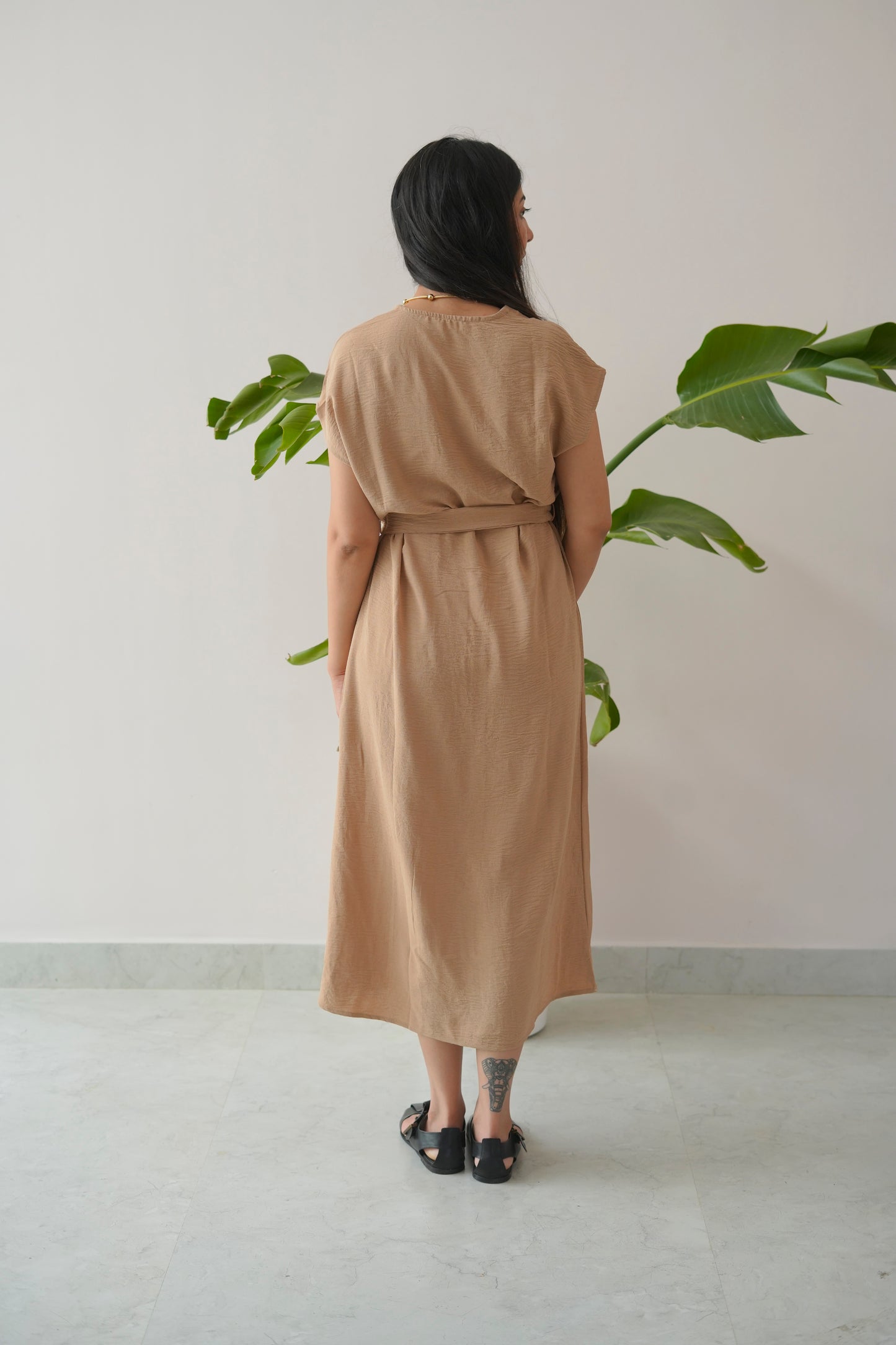 Woman wearing a beige dress standing in front of a plant against a plain wall.