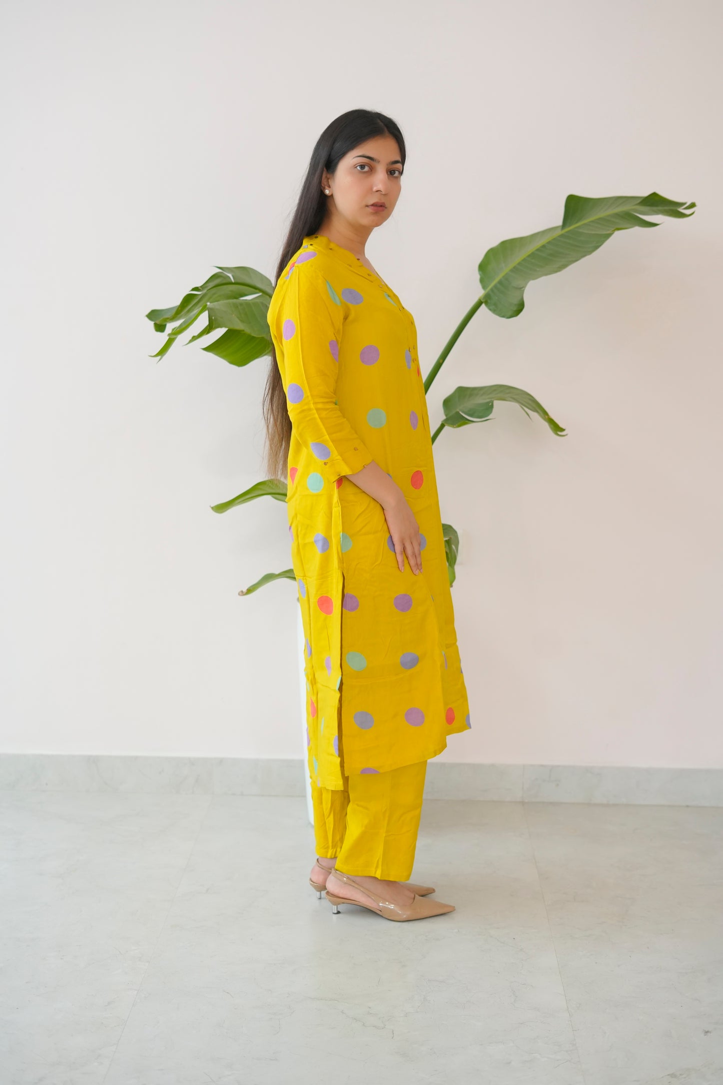 Woman wearing a yellow outfit with colorful polka dots standing next to a plant against a white wall.