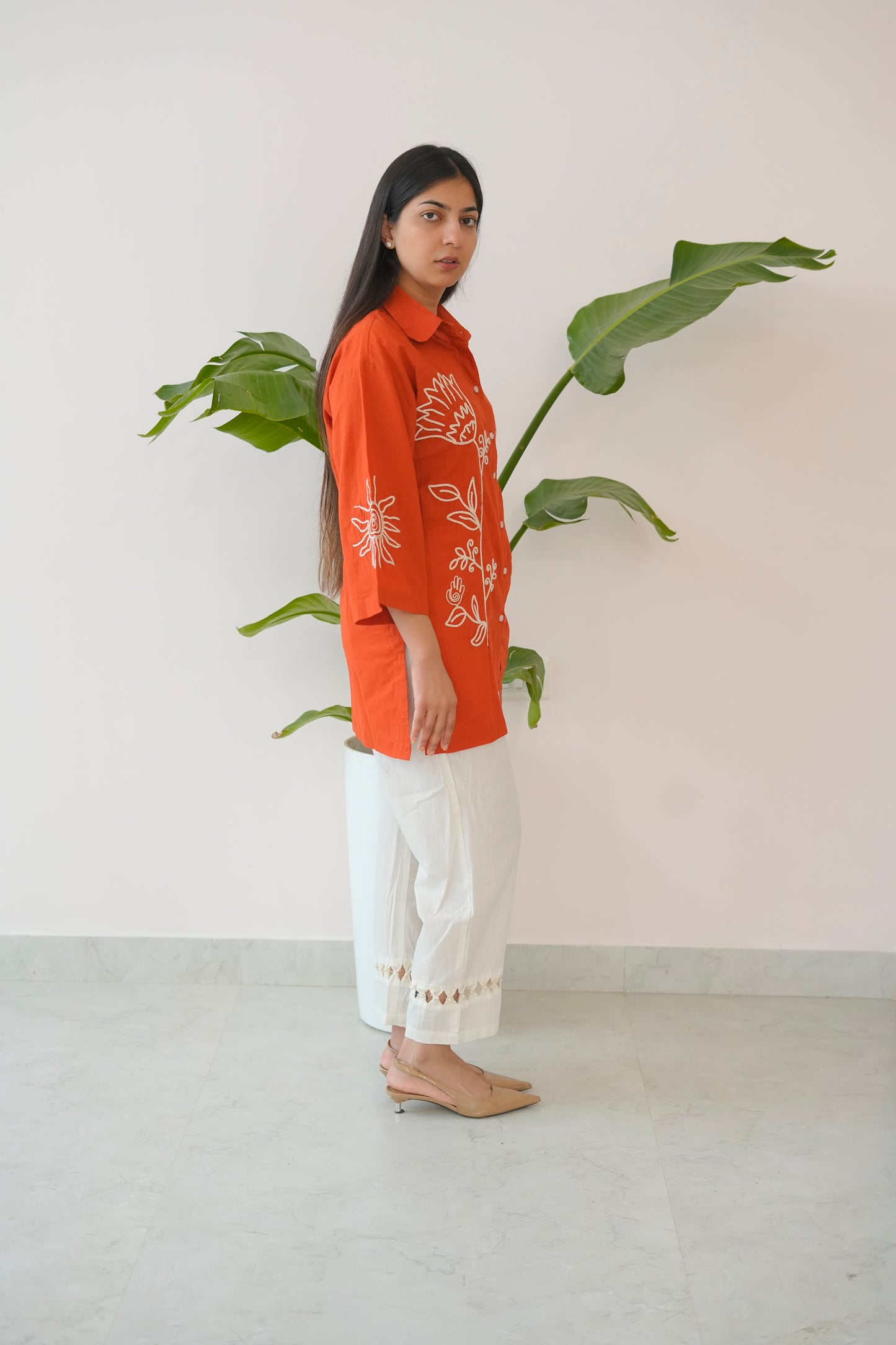 Woman wearing an orange embroidered shirt and white pants standing next to a plant against a plain background