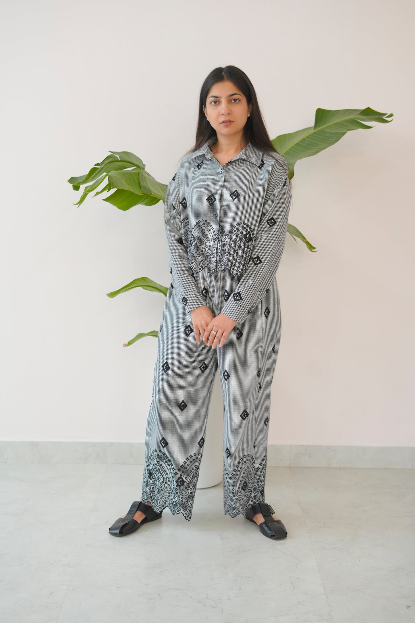 Woman wearing a gray patterned outfit standing against a white wall with green leaves.