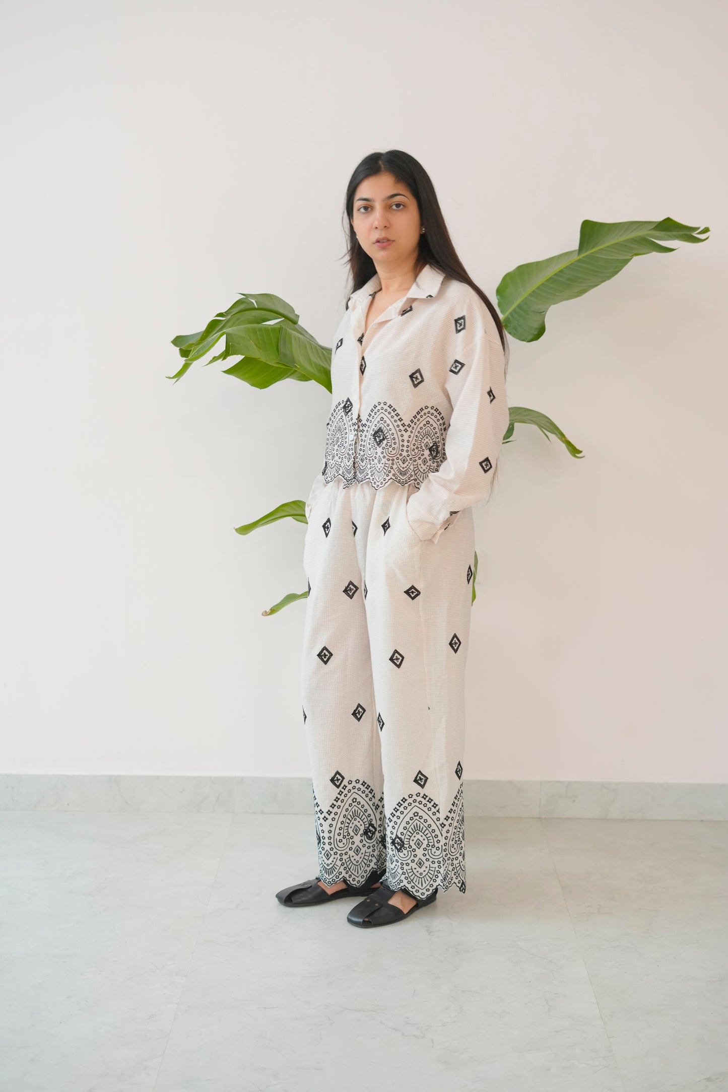 Woman wearing a white outfit with black patterns standing against a white wall with green leaves.
