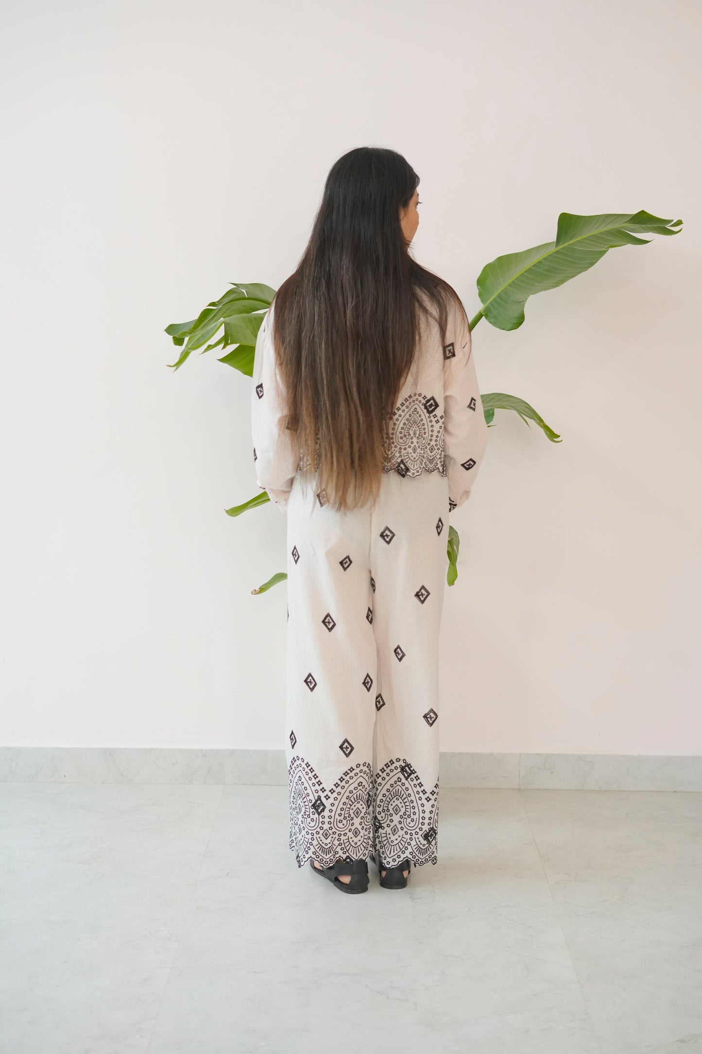 Woman wearing a long, light-colored dress with black patterns standing against a white wall with green plants.