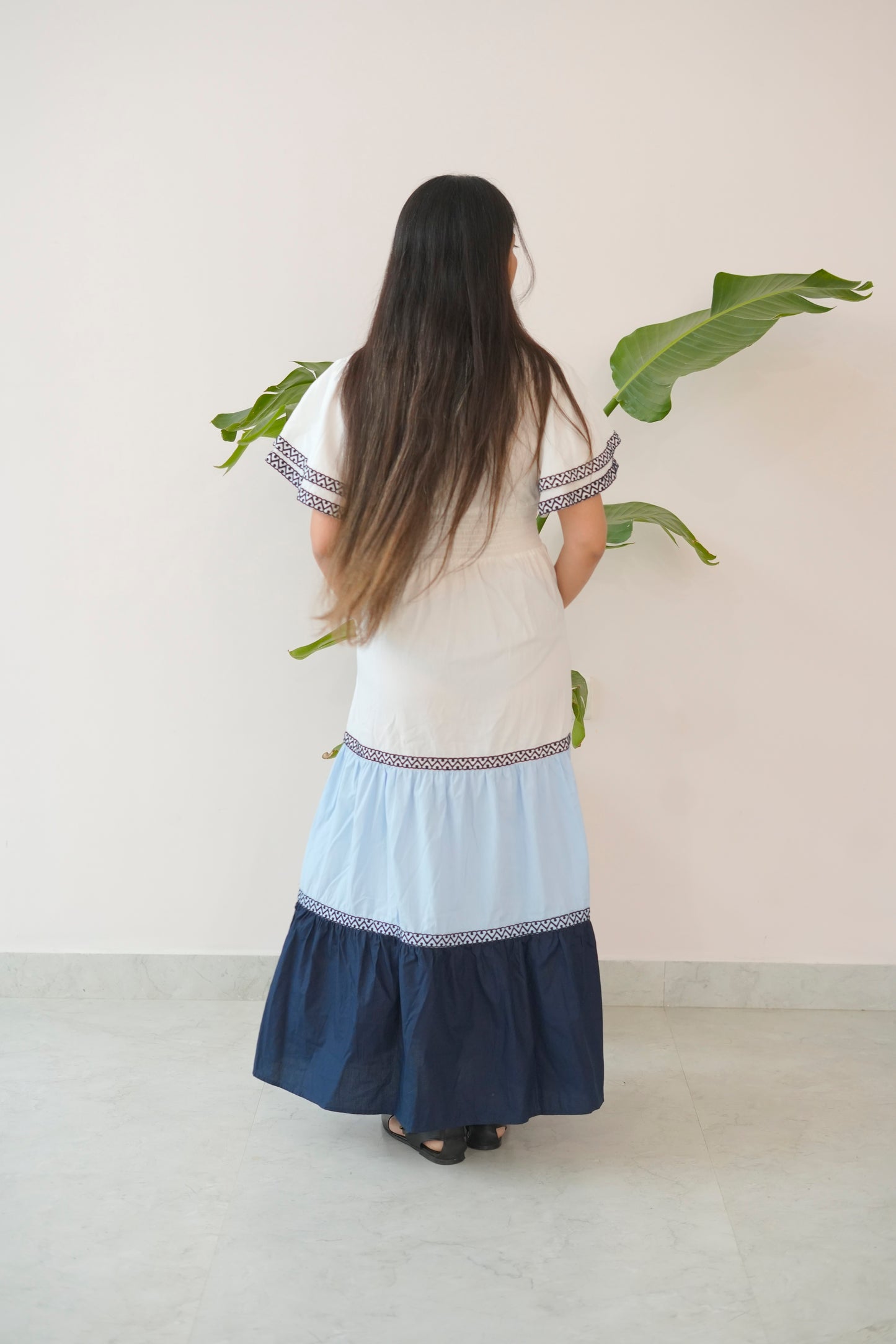 Woman wearing a white top and blue skirt with embroidery, standing against a light background with green leaves.