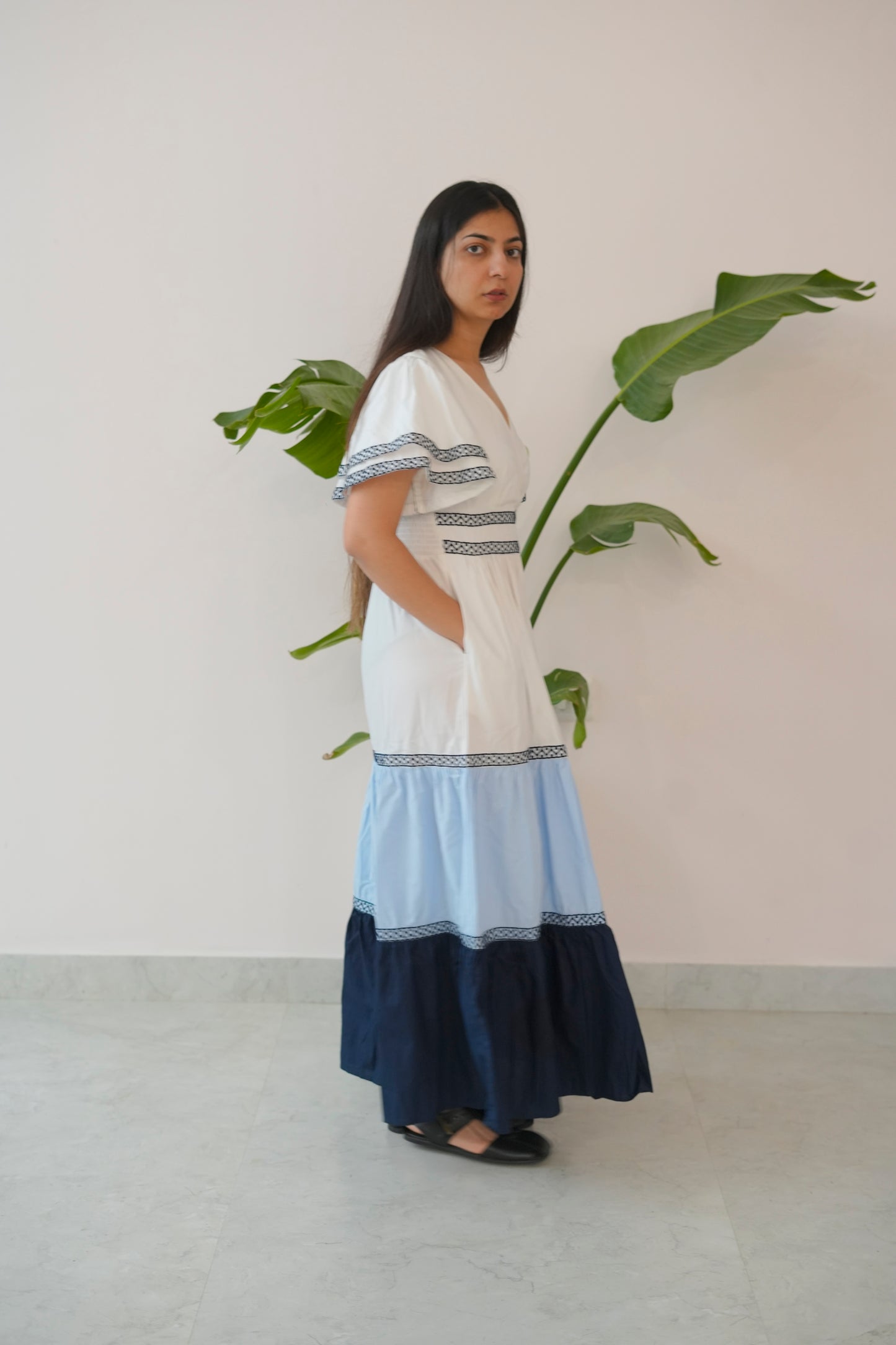 Woman wearing a white dress with blue accents standing next to a plant against a plain wall.