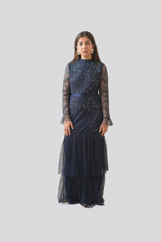 Woman wearing a dark blue dress with sheer sleeves against a white wall with green leaves.