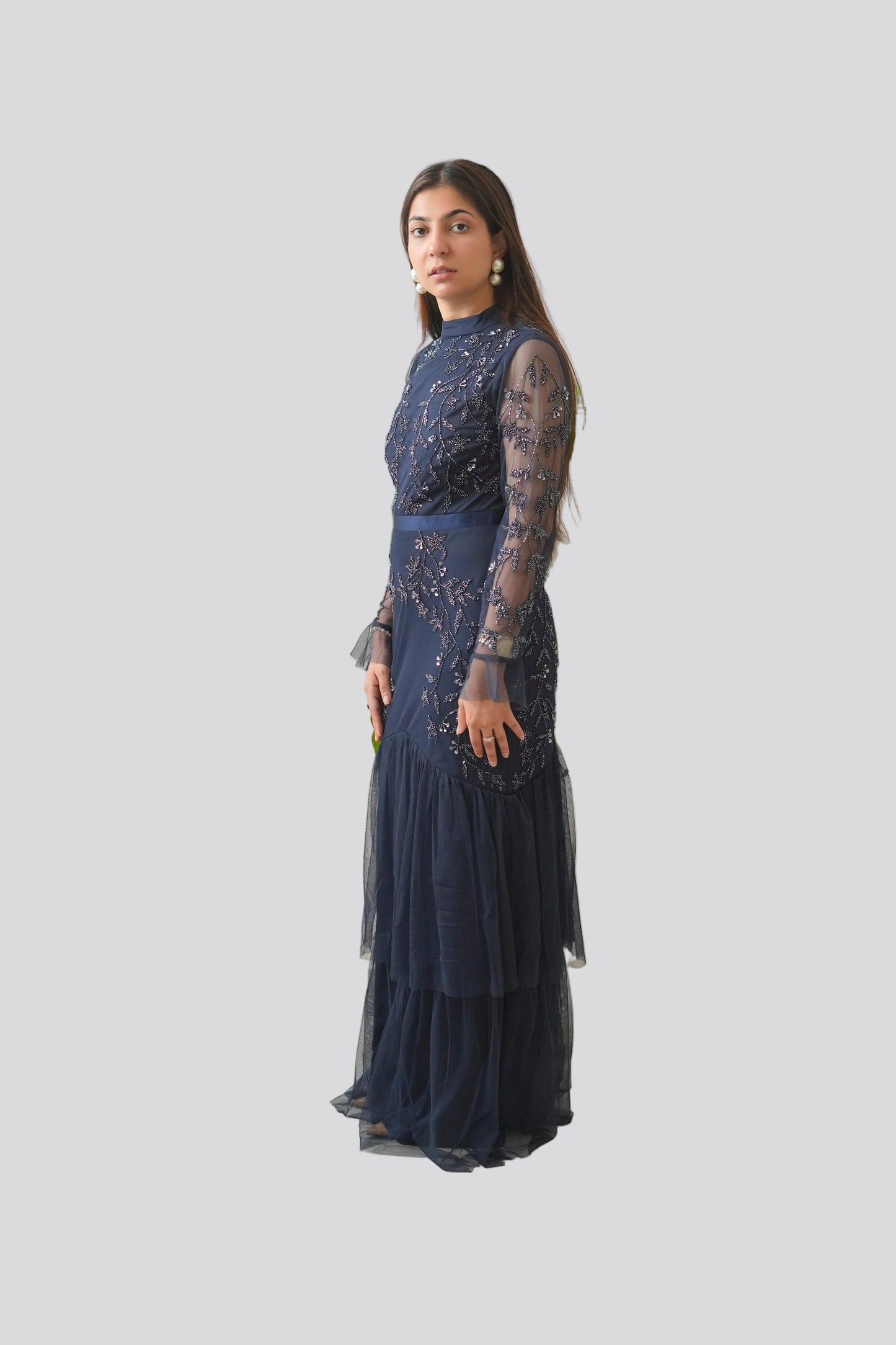 Woman in a dark blue embroidered dress standing against a white wall with green plants.