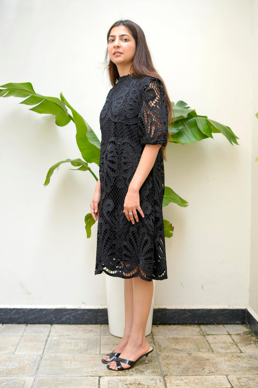 Woman wearing a black lace dress standing against a white wall with green plants.