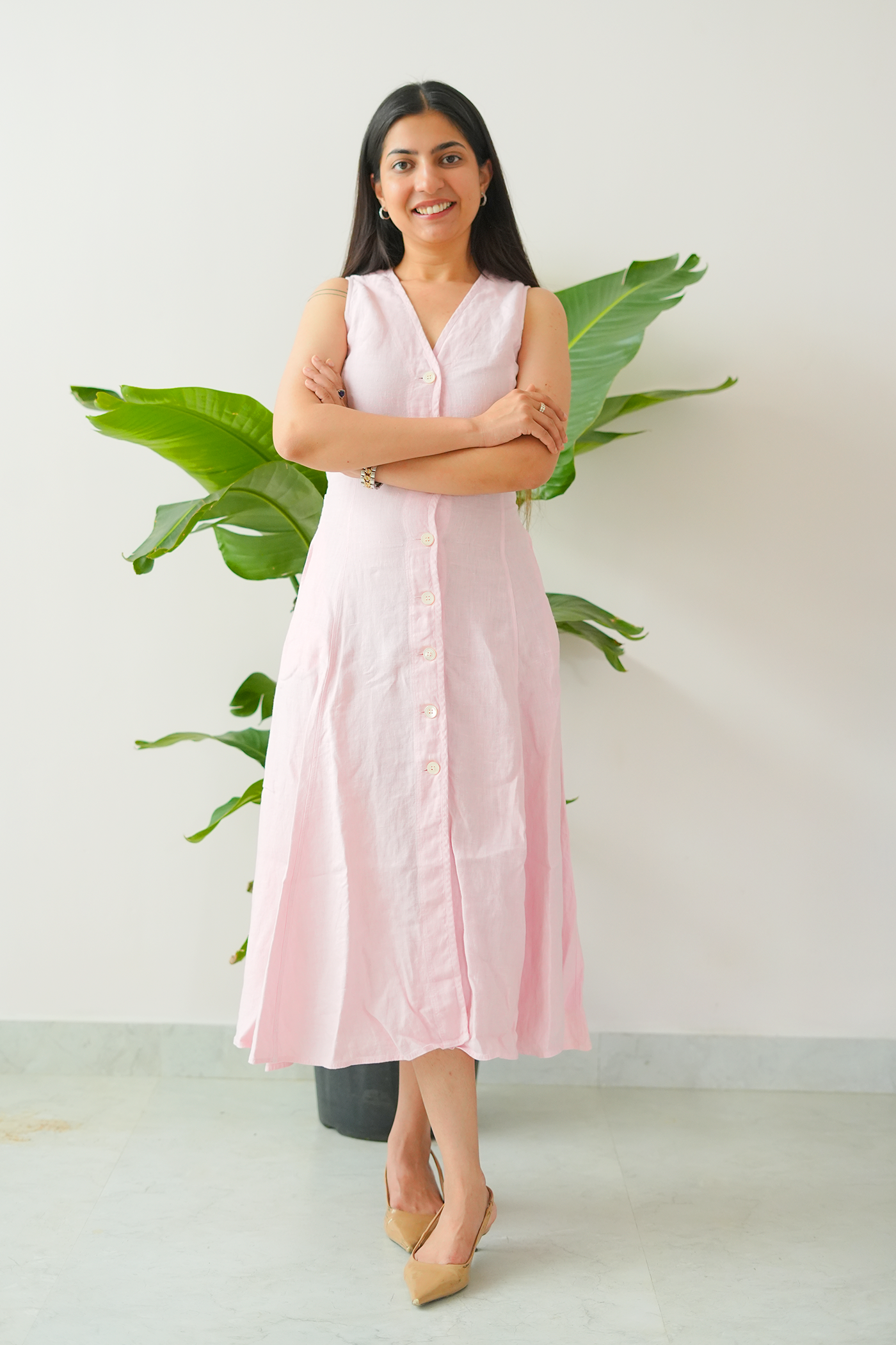 Woman wearing a light pink sleeveless dress standing in front of a plant against a white wall.