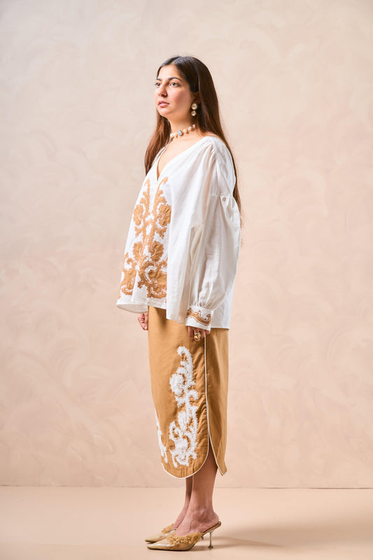 Woman wearing a white blouse with gold embroidery and beige pants against a beige background