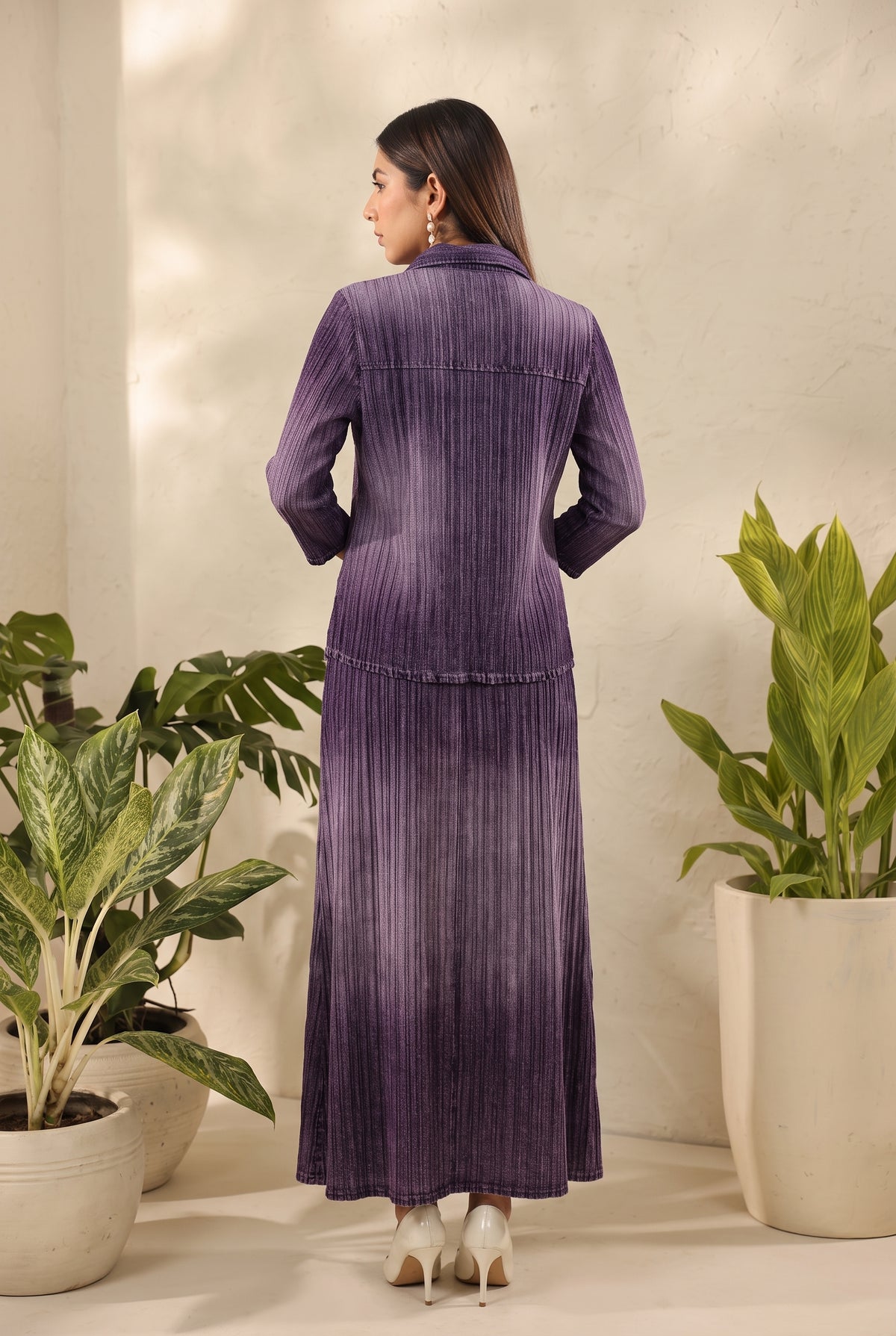 A woman stands with her back to the camera wearing a long-sleeved, high-neck purple pleated maxi dress. She is posed in a studio setting with potted green plants on either side against a textured off-white wall.