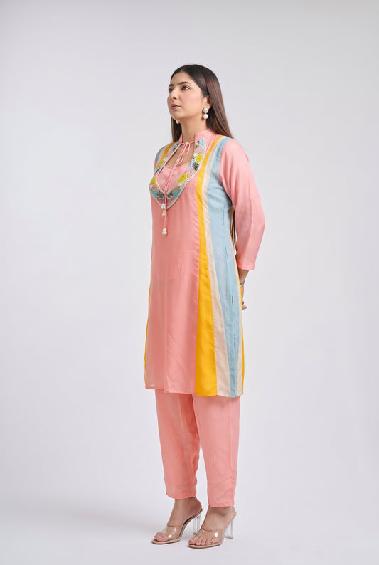 A woman modeling a light pink kurta set with pastel blue and yellow side panels, a colorful embroidered neckline, matching pink pants, and clear high-heeled sandals, posed against a light gray studio background.