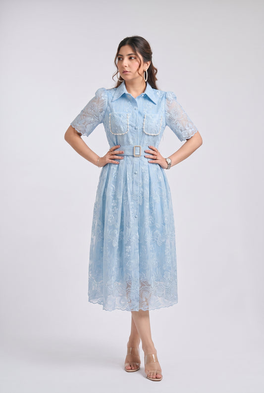 A woman modeling a light blue midi dress with sheer lace short sleeves, a button-up front, decorative chest pockets with pearl trim, and a matching lace belt with a gold buckle, set against a plain light gray studio background.