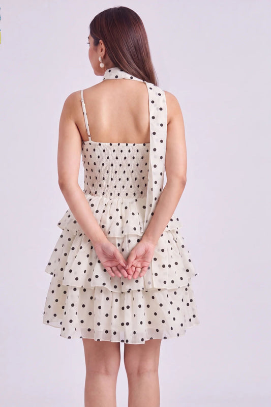 A woman shown from the back wearing a sleeveless halter-neck dress with a tiered ruffle skirt in a warm off-white fabric with black polka dots, set against a plain light gray studio background.