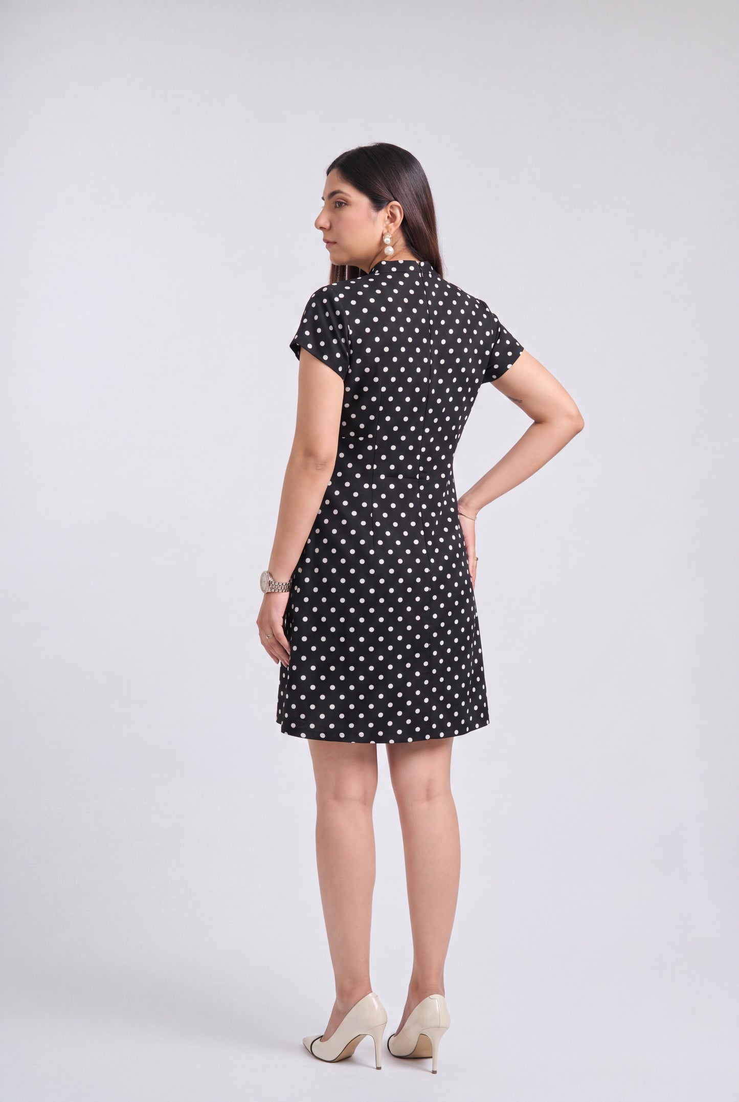 A woman modeling a short-sleeve black dress with a white polka dot pattern, shown from a rear three-quarter view against a light gray studio background.