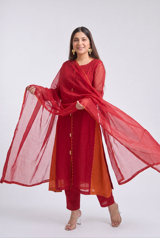 A woman modeling a red three-piece traditional outfit with a matching sheer dupatta, featuring a red kurta with gold button accents, red pants, and a rust-orange inner lining, set against a plain light gray studio background.