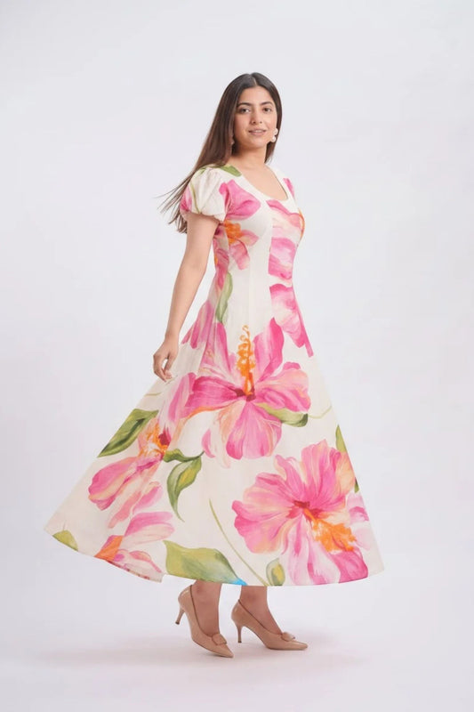A woman modeling a short-sleeve V-neck maxi dress with a vibrant pink and orange hibiscus floral print on a cream background, set against a plain light gray studio backdrop.