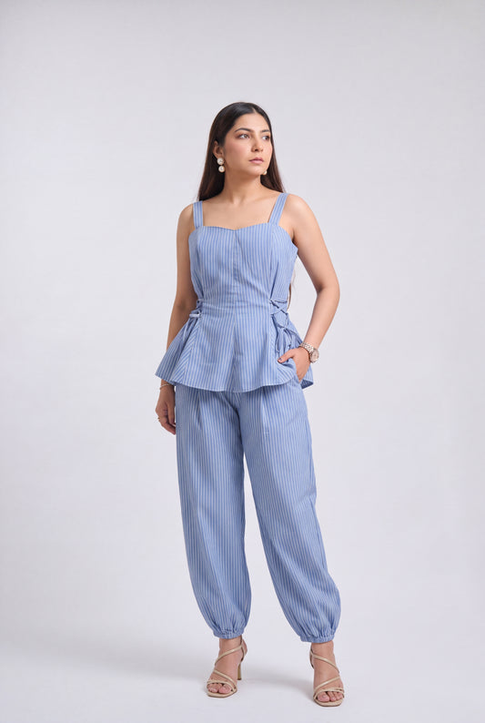 A woman modeling a light blue and white vertically striped two-piece set consisting of a sleeveless peplum top and matching wide-leg pants, photographed against a neutral light gray studio background.
