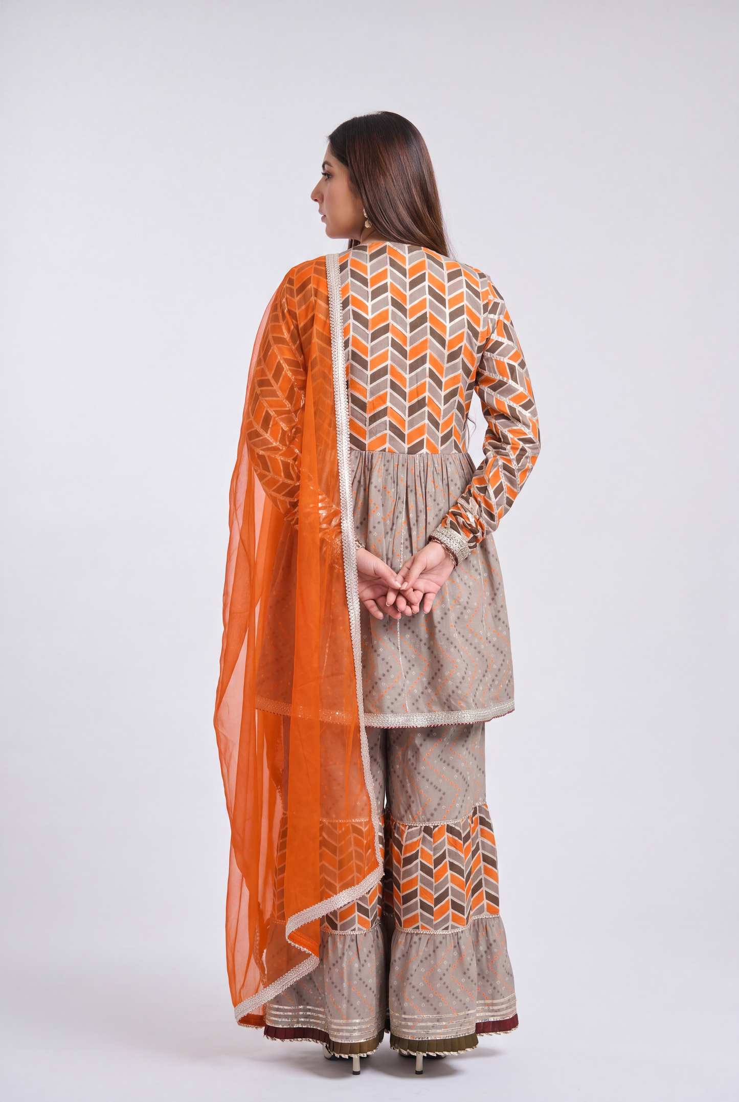 A woman modeling a traditional South Asian outfit consisting of a beige kurta and wide-leg pants with orange and brown geometric patterns, paired with a bright orange dupatta, against a light gray studio background.