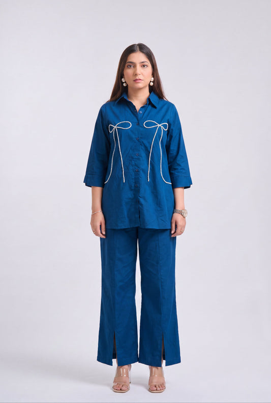 A woman modeling a matching blue two-piece set consisting of a long-sleeve button-up shirt with white bow embroidery and matching wide-leg trousers, shot against a light gray studio background.